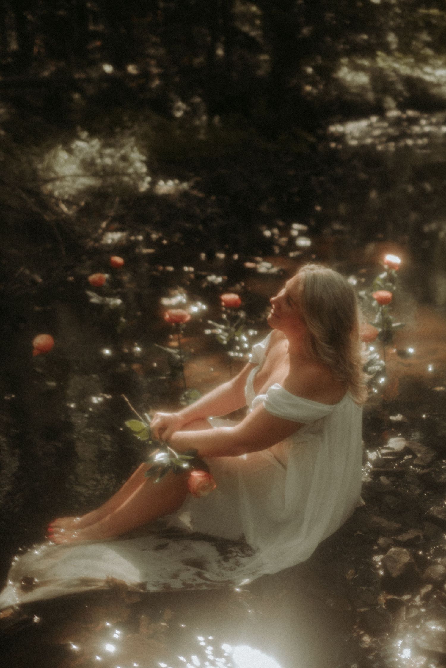 An ethereal figure in white floats gracefully in dark woodland waters surrounded by scattered flower petals.