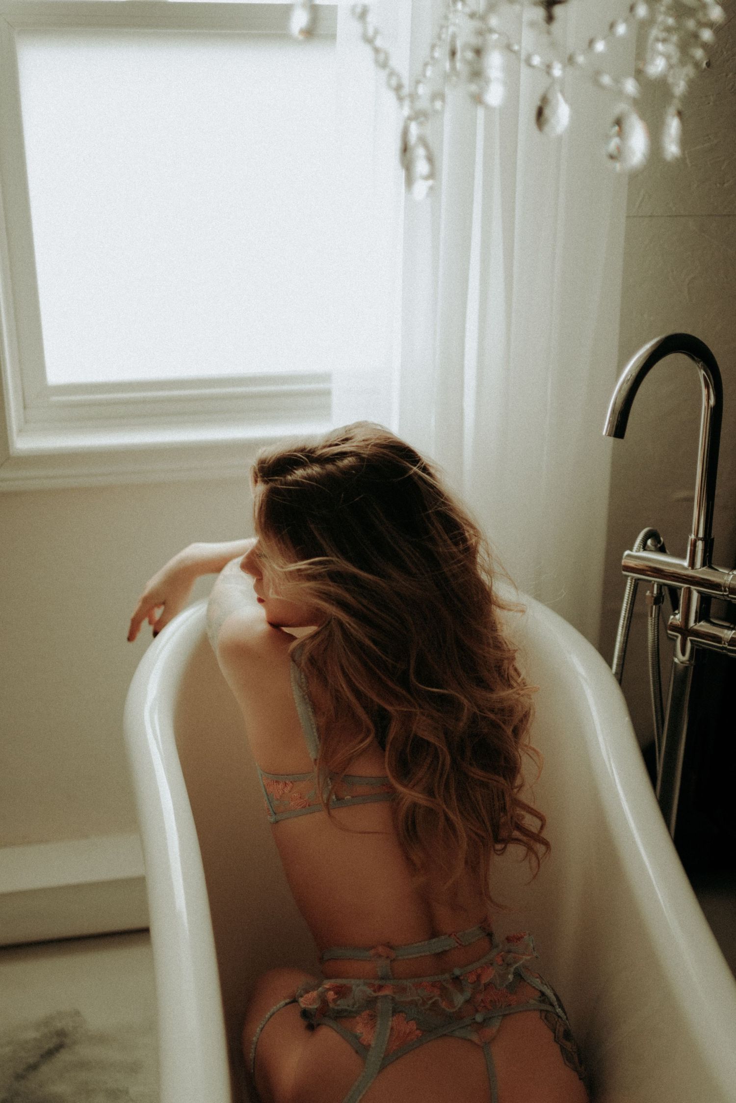 Person with wet curly hair leaning back in a white bathtub with natural light in vintage style bathroom settings.
