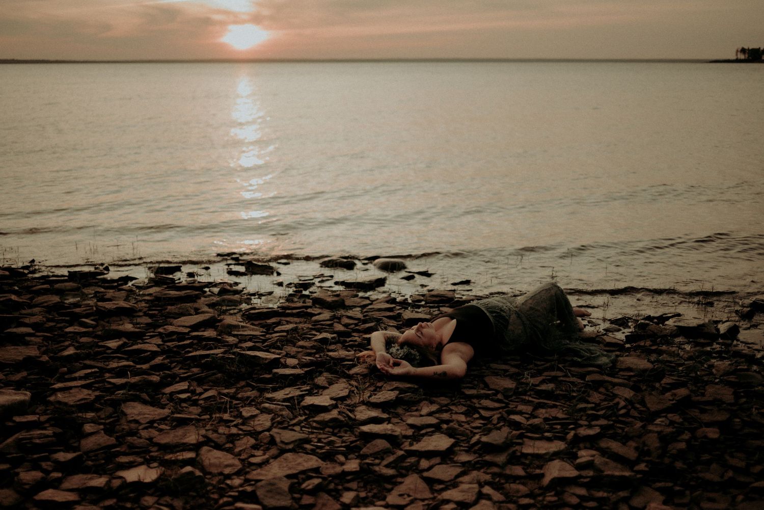 Person lying on rocky beach at sunset with water and horizon in background.