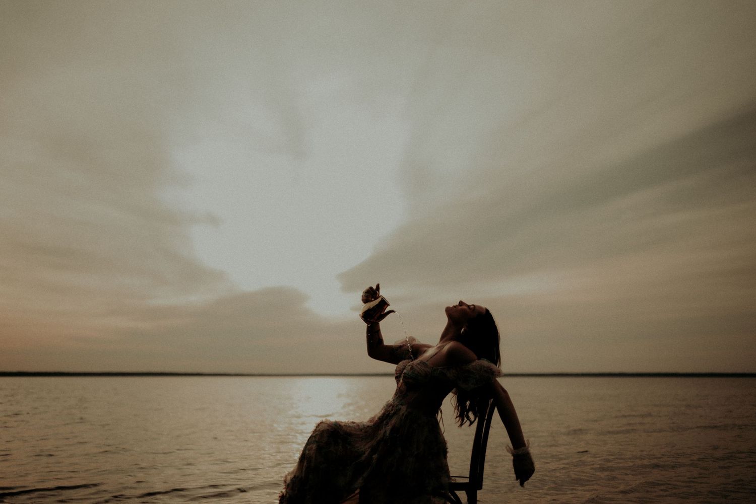 Silhouette of a figure in a dress posing on a chair against a serene lake at dusk.