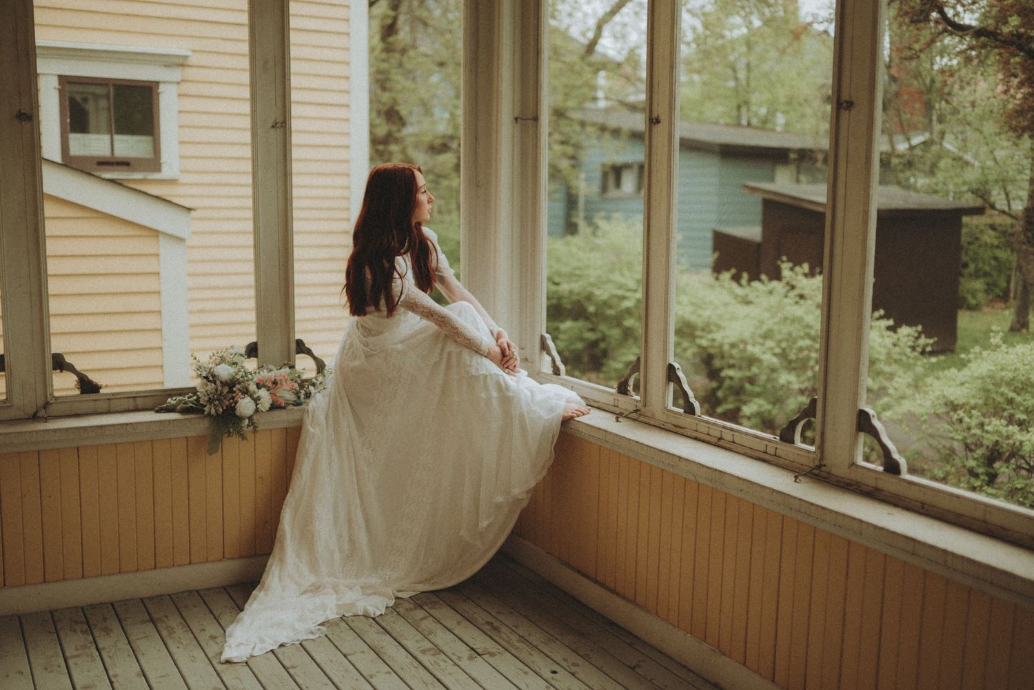 A rustic wooden porch with railing wraps around a light yellow house surrounded by trees on a cloudy day.