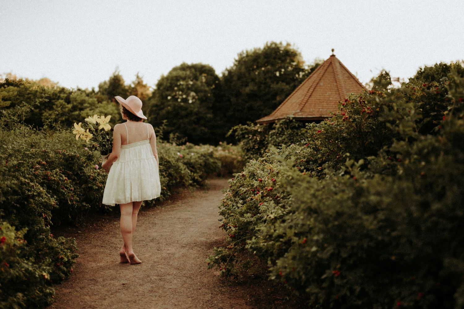 A person in a white dress walks through a lush garden path surrounded by blooming flowers and greenery at sunset.