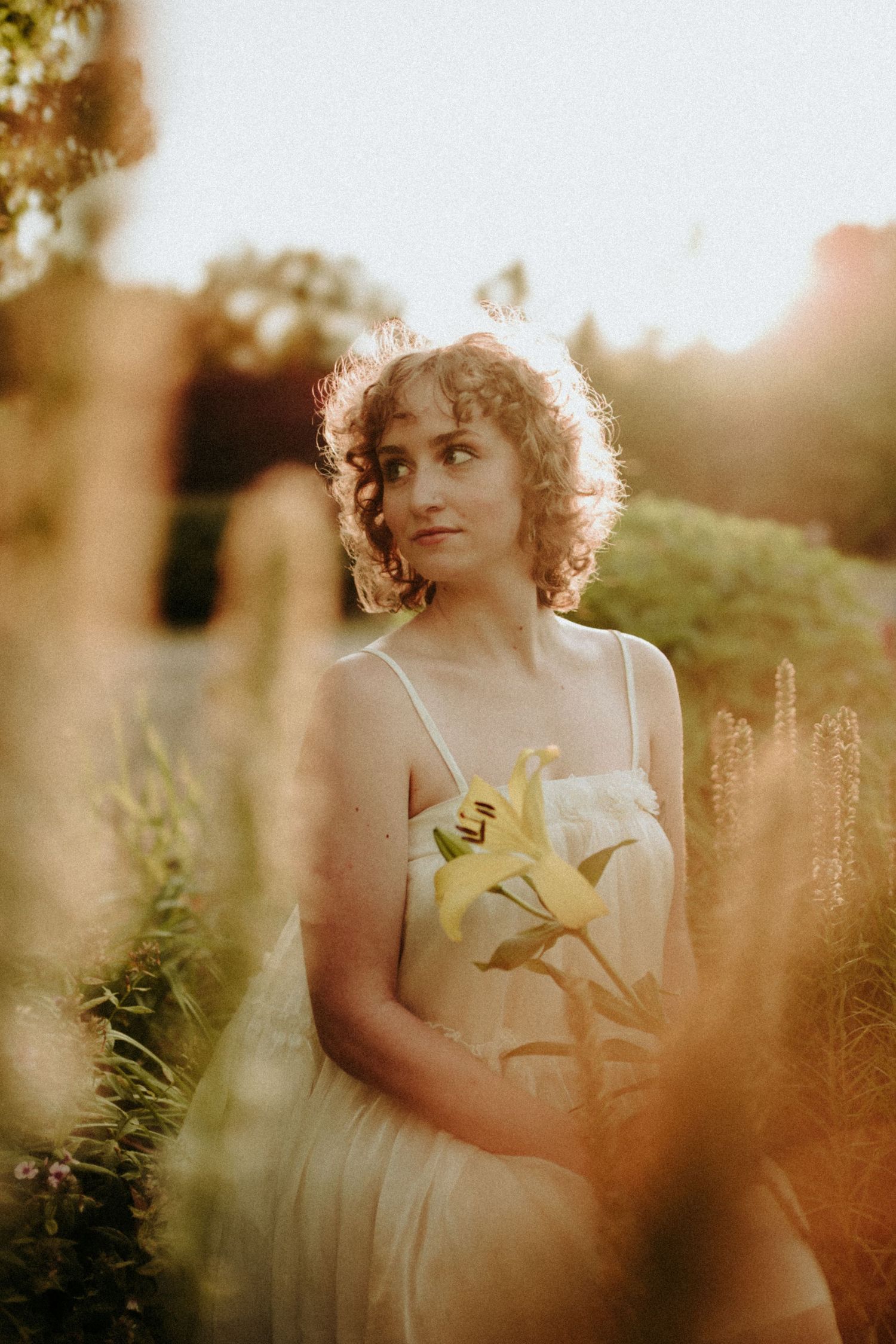 A romantic sunset photo showing someone in a white sundress holding flowers while surrounded by garden foliage.