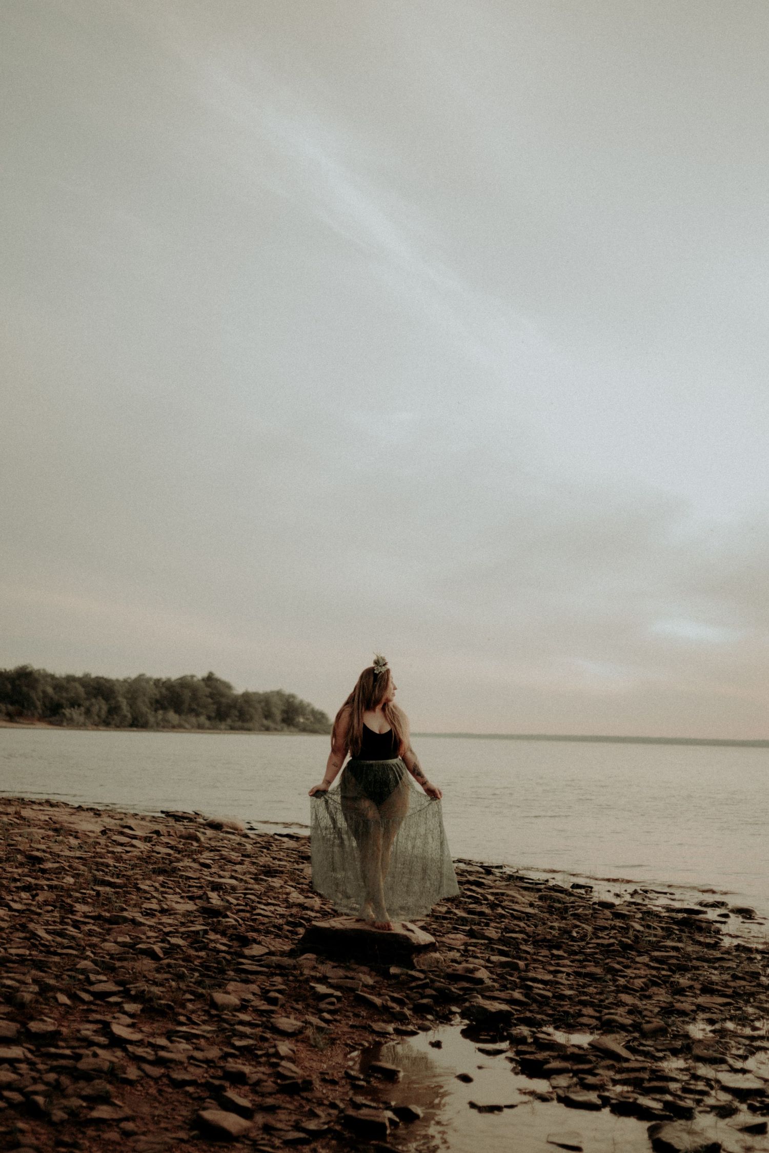 A figure in a flowing dress stands on a rocky shoreline at dusk overlooking a serene lake.
