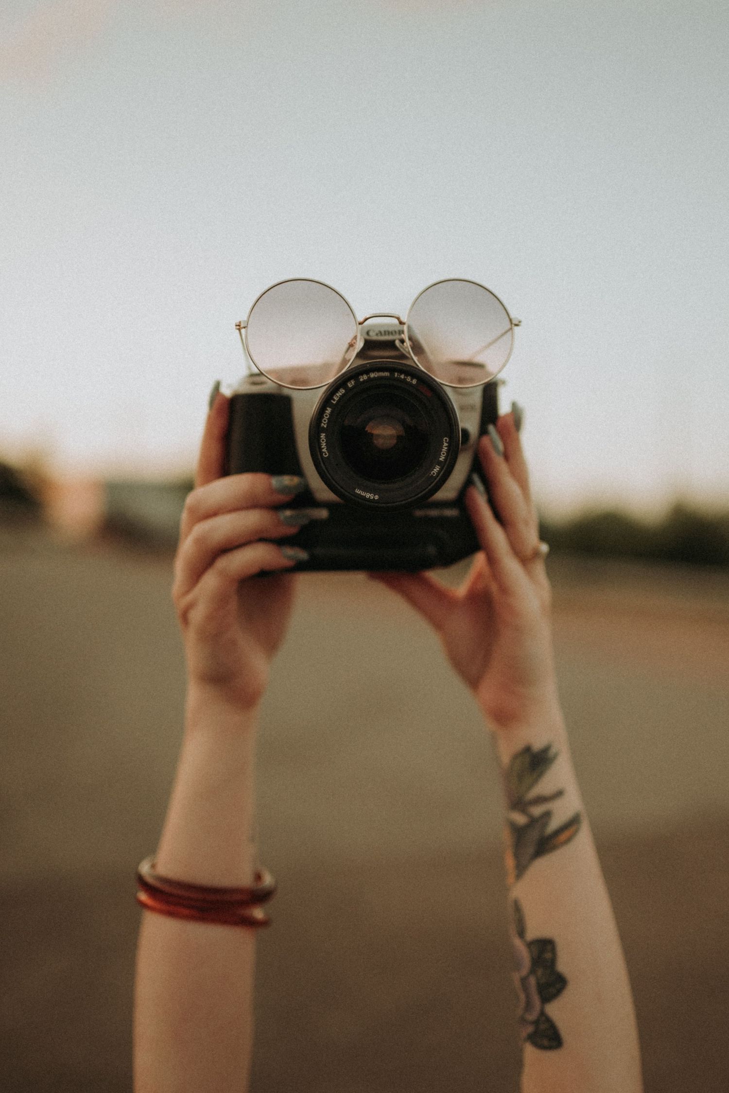 Hands holding vintage camera with round sunglasses perched on top against blurred background.