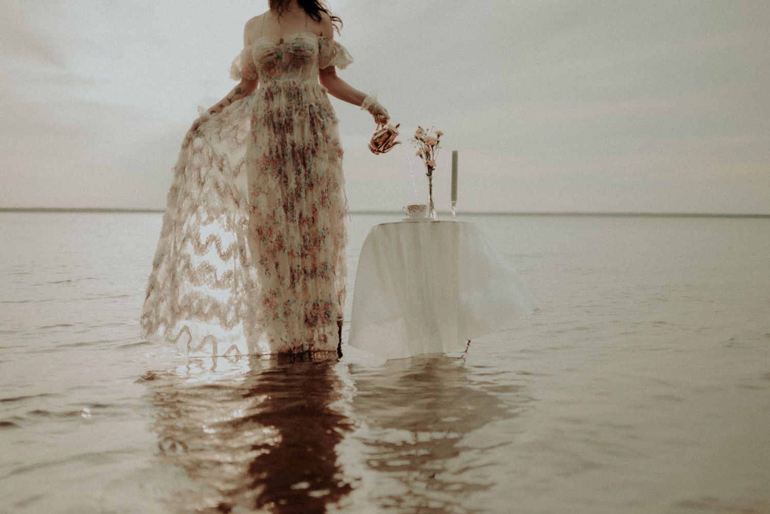 Artistic water portrait with flowing dress and vintage table setup in sepia tones.