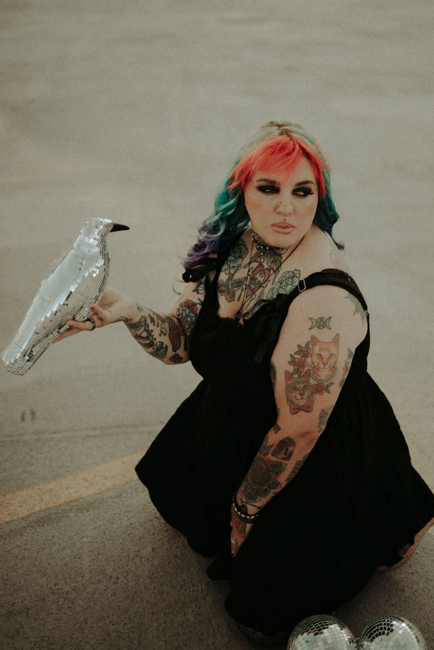 Alternative model in black dress with colorful hair poses with white bird on beach at sunset.