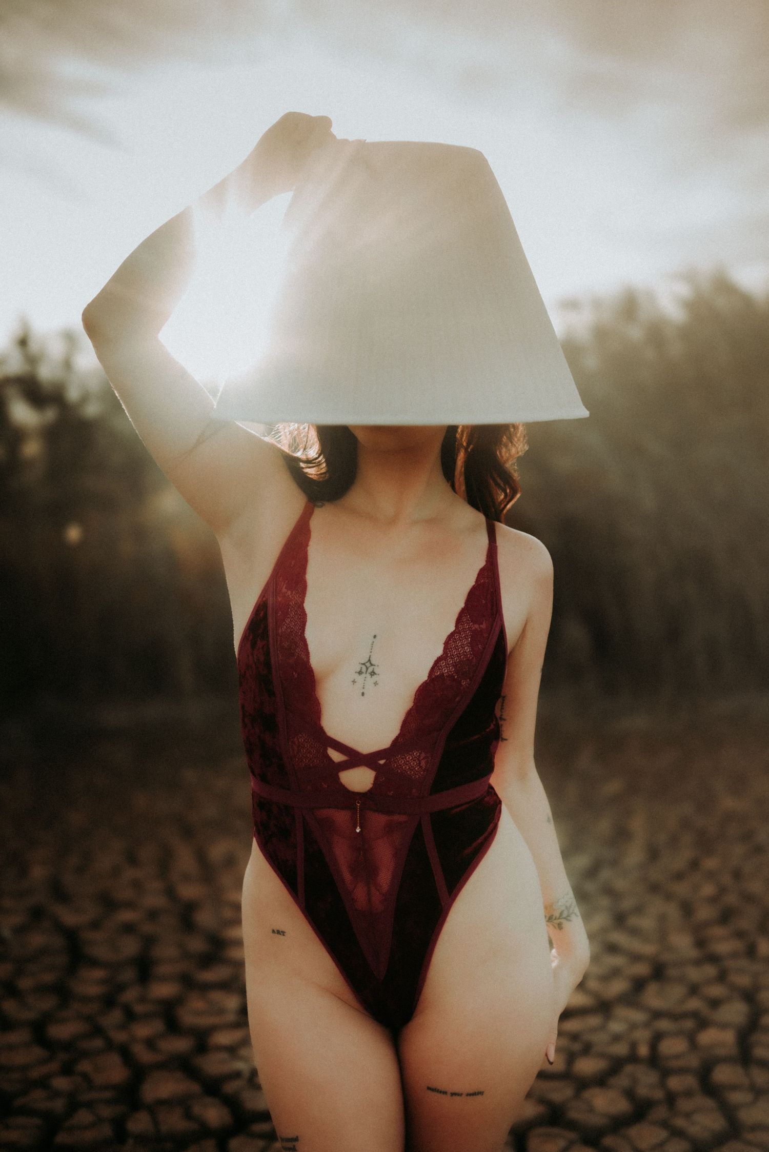 A person wearing a burgundy bodysuit poses in front of a sun-drenched cracked earth landscape at sunset.
