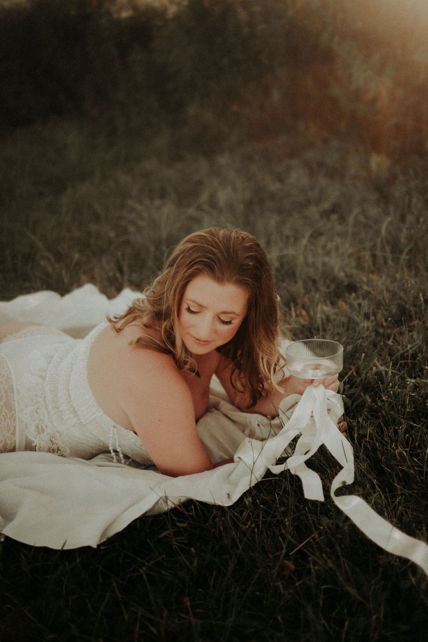 Ethereal portrait of someone lying in white fabric on grass at sunset with dreamy lighting.