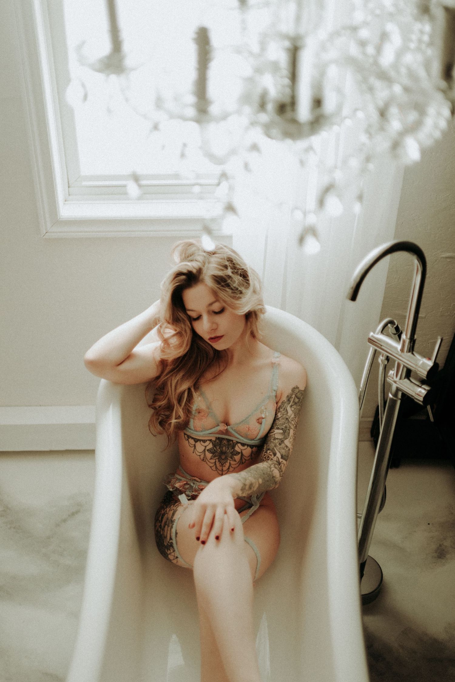 Artistic boudoir photograph of woman in vintage clawfoot bathtub with window light casting soft shadows.