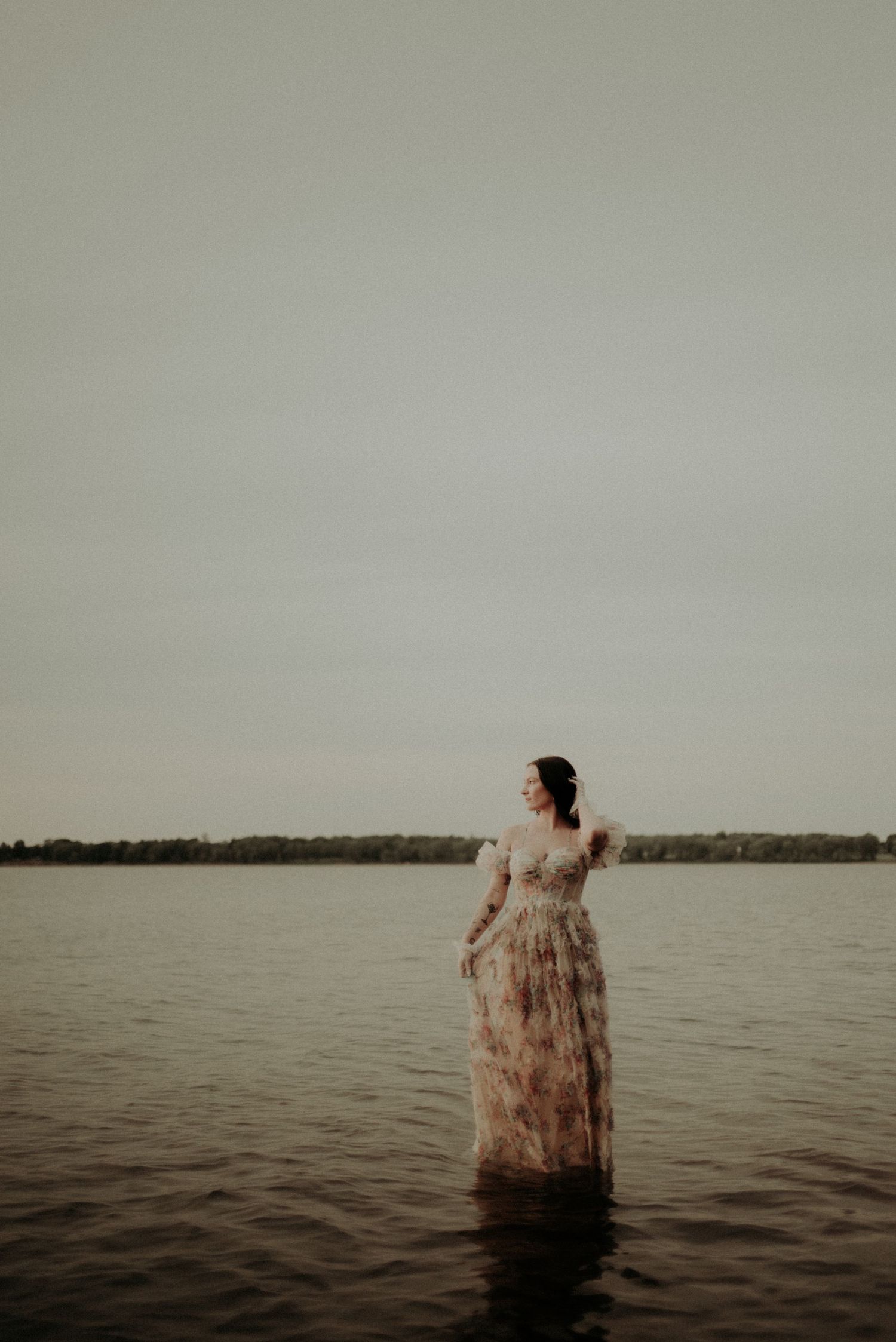 A figure in a floral dress stands in tranquil waters at dusk, creating a dreamy atmospheric scene.