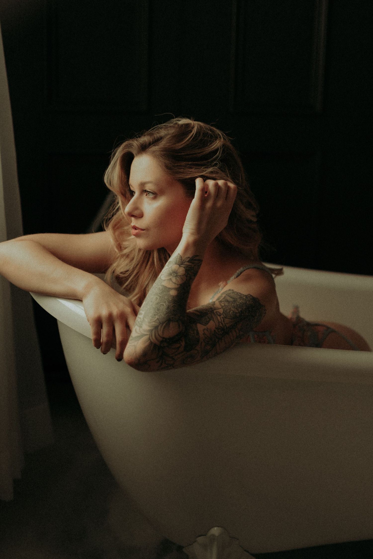Moody boudoir photo of person with tattoos relaxing in white bathtub against dark background.