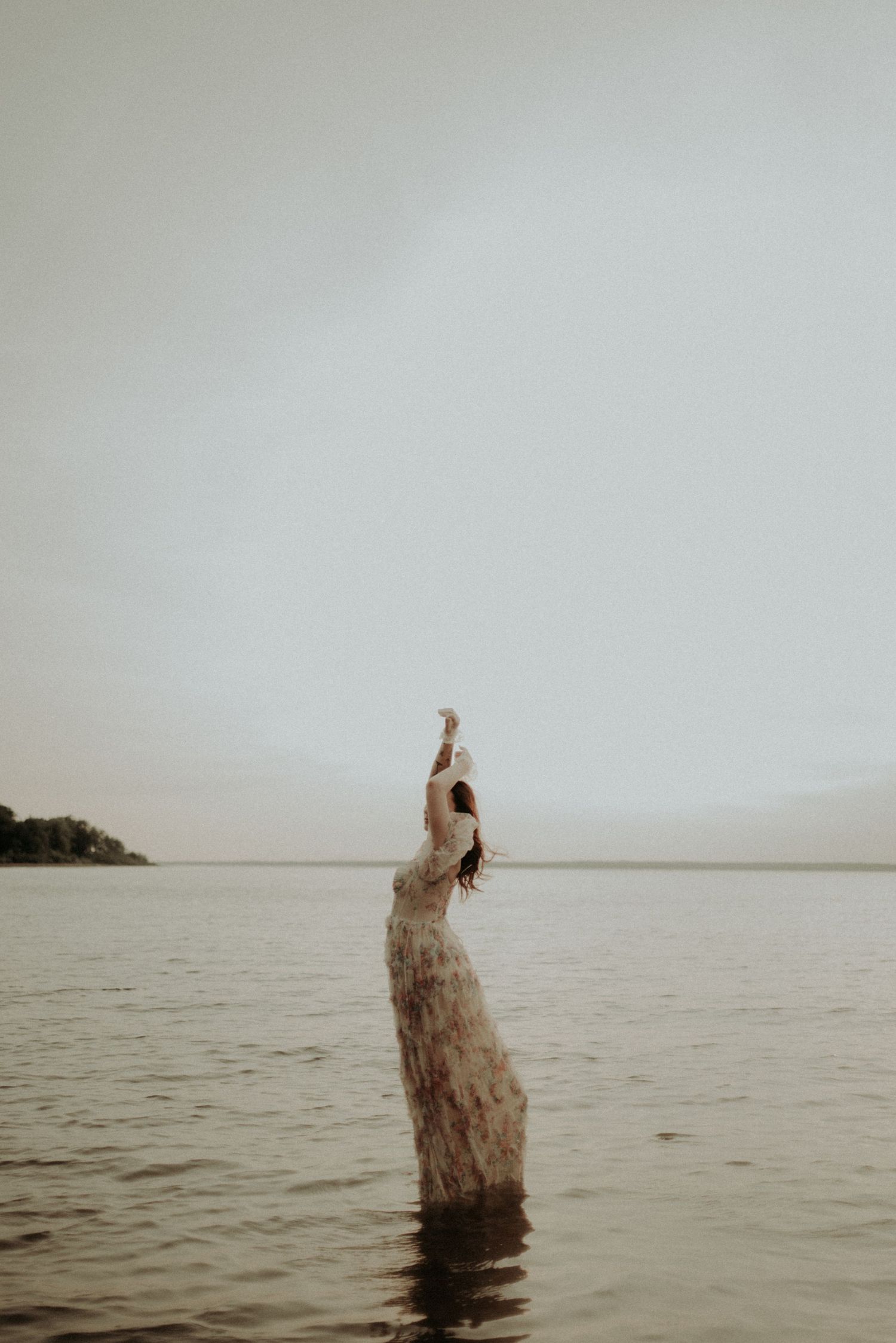A graceful pose with arms raised in shallow waters wearing a flowing dress.