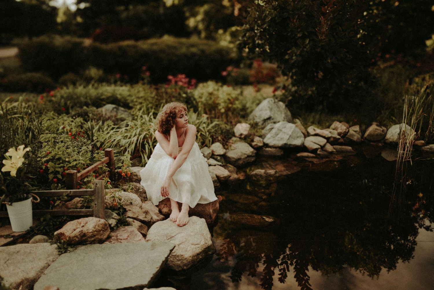Figure in a flowing white dress sits pensively by a garden pond surrounded by rocks and greenery in warm sunlight.