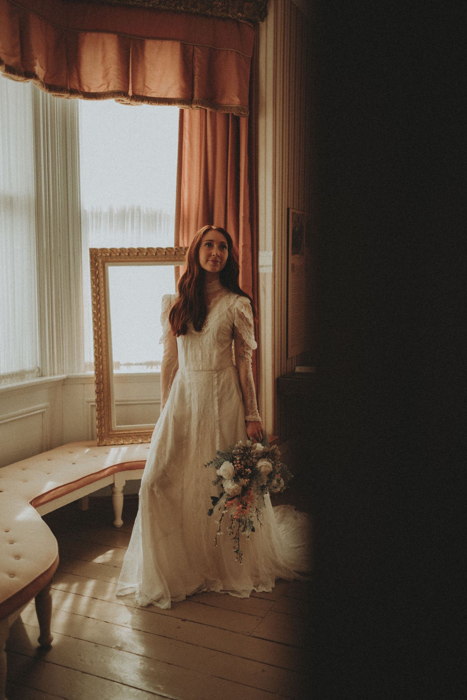 Moody wedding photography in a sunlit room with flowing white gown and fresh floral bouquet.