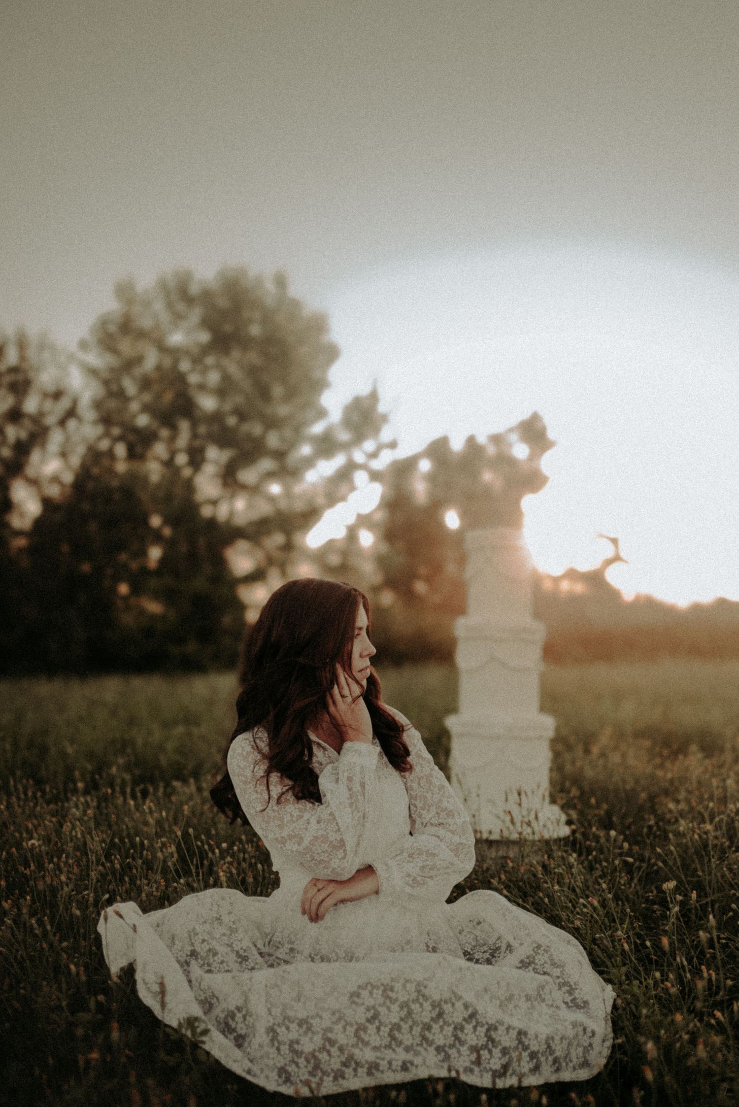 A person in a flowing white lace dress sits gracefully in a sunlit meadow at golden hour.