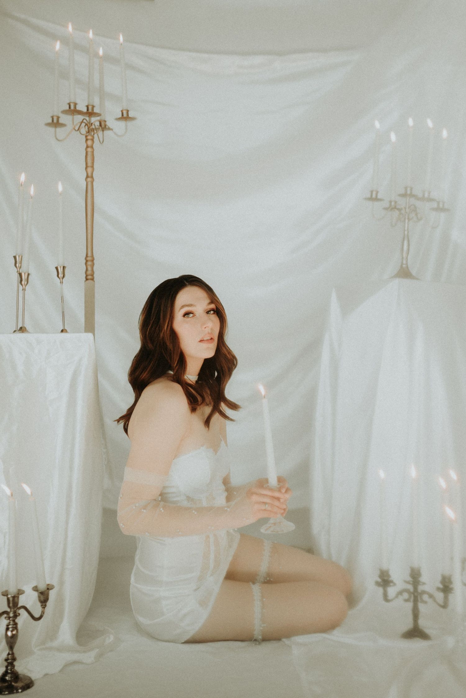 Ethereal boudoir photoshoot in white draped room with vintage candelabras and soft lighting creates dreamy atmosphere.