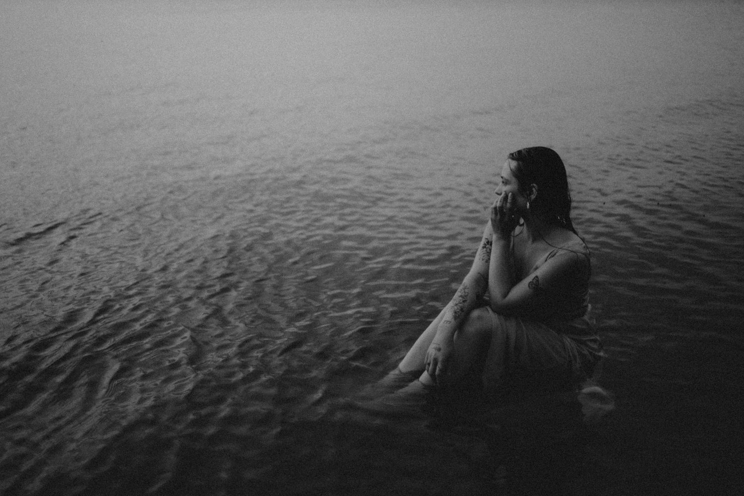 Moody monochrome image of someone sitting in water against a serene lake backdrop.