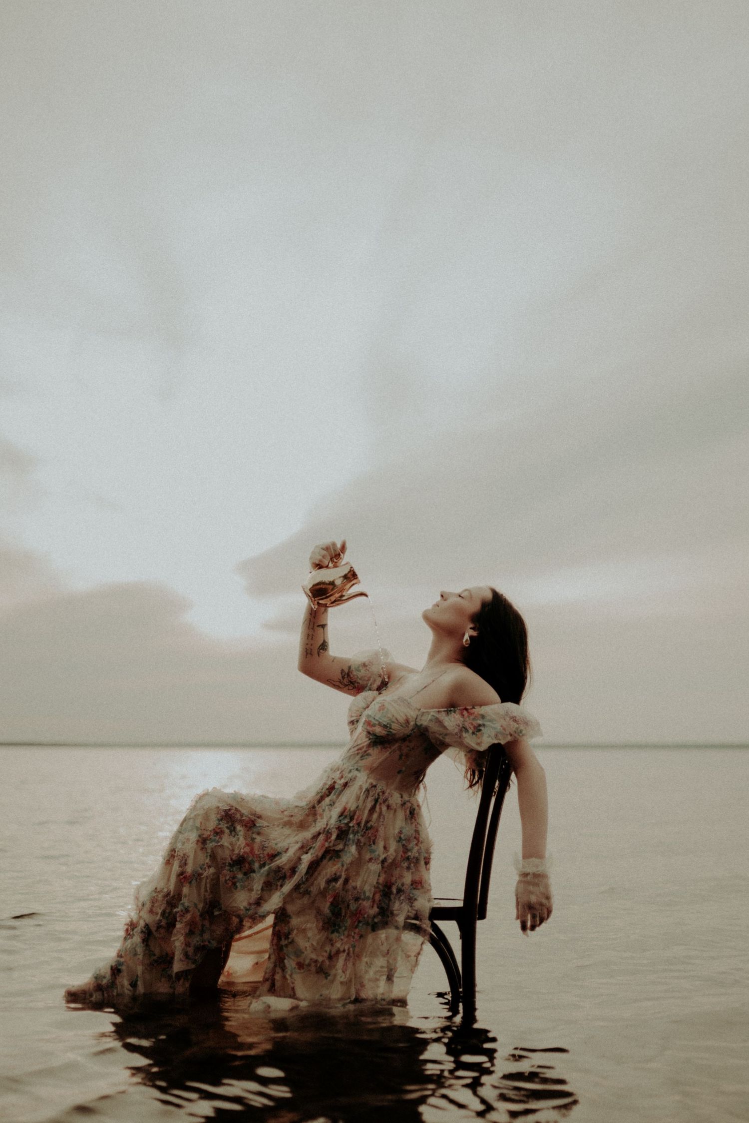Artistic portrait of a person in a flowing dress reclining on a chair in shallow water at sunset.