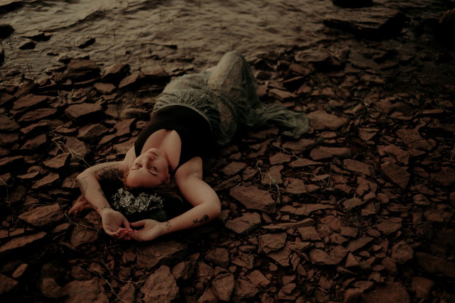Figure reclining on rocky shoreline during moody sunset photoshoot.