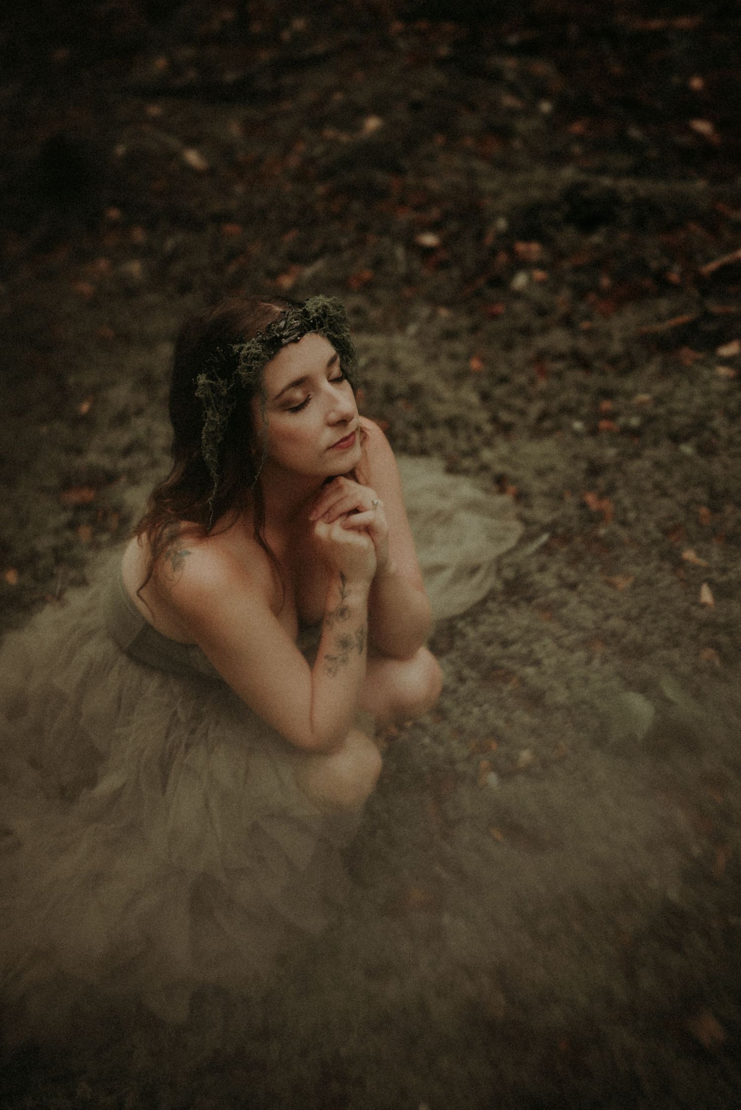 Dreamy portrait in a tulle gown and floral crown against moody forest backdrop with soft lighting and earthy tones.
