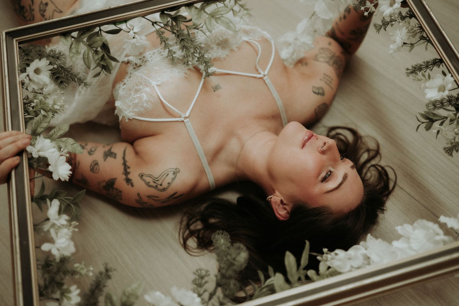 Artistic boudoir photograph with white lace lingerie and floral elements in soft, moody lighting.
