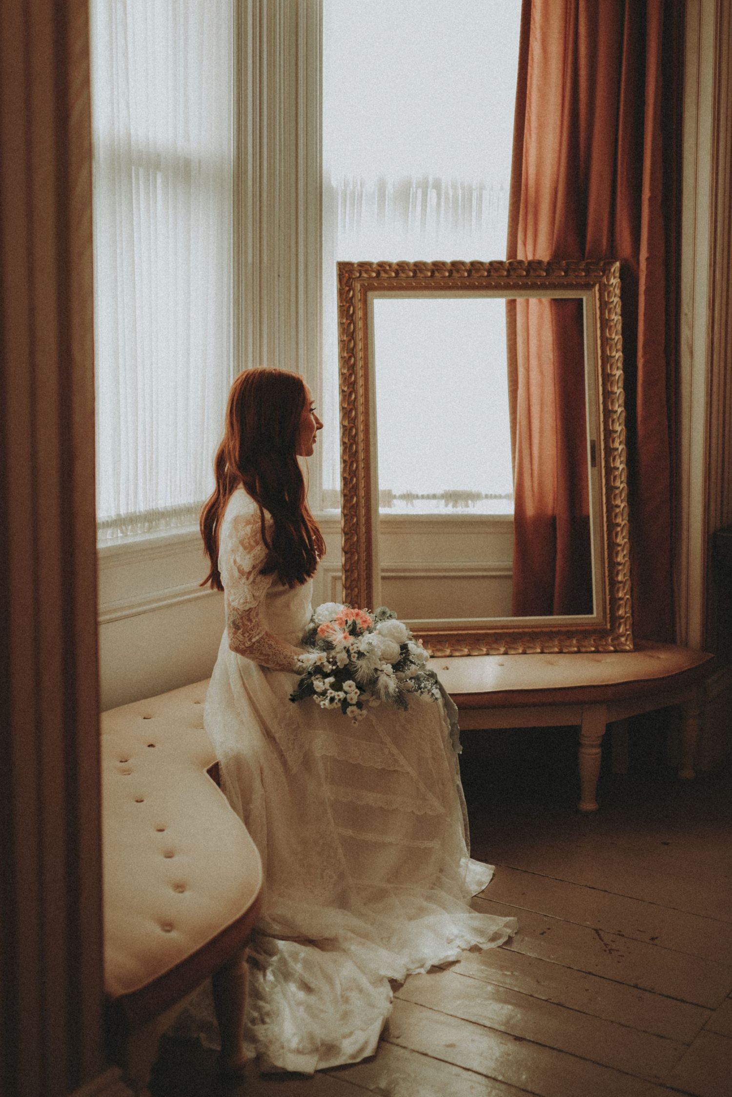 A vintage-style scene showing someone in a flowing white wedding dress sitting by sunlit windows with ornate gold frames.