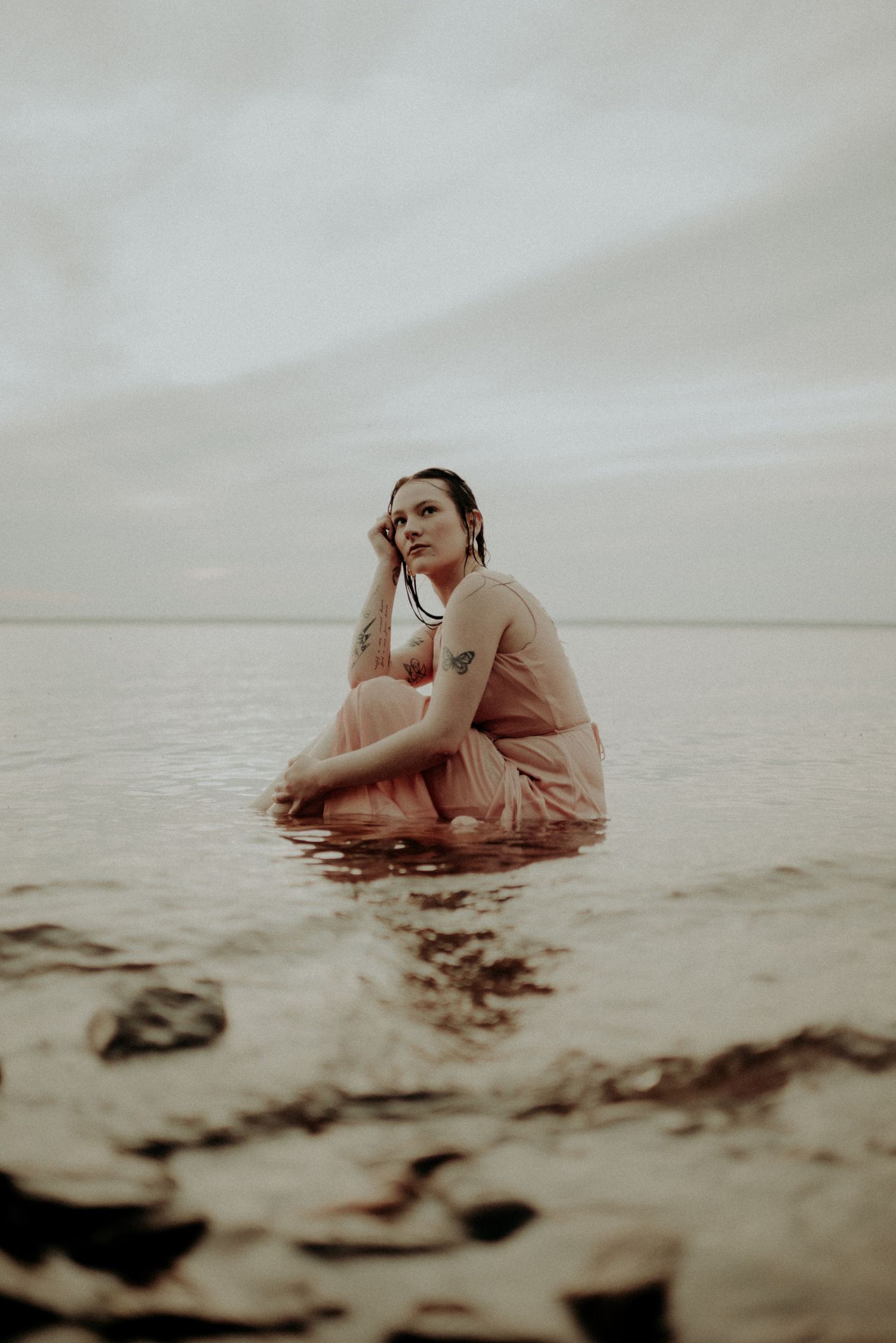 A person in muted tones sits quietly in shallow coastal waters.