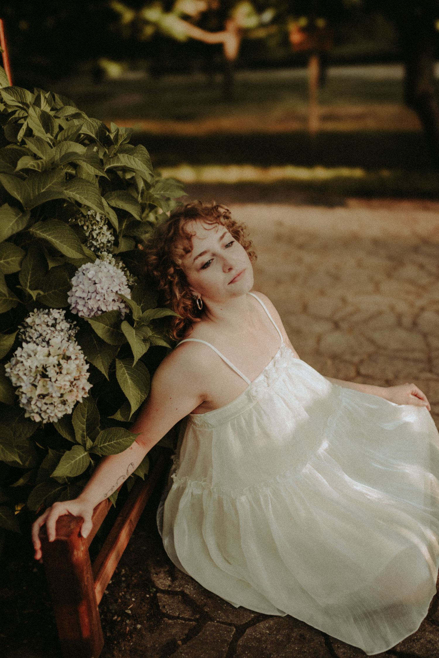 Dreamy portrait of a person in a white dress reclining near blooming hydrangea flowers in a garden setting.