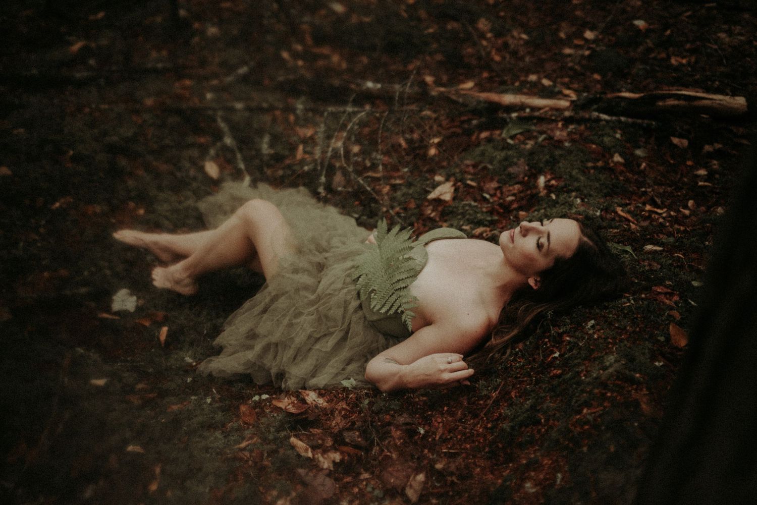 A figure lies peacefully on a bed of autumn leaves in the forest during a moody outdoor photoshoot.