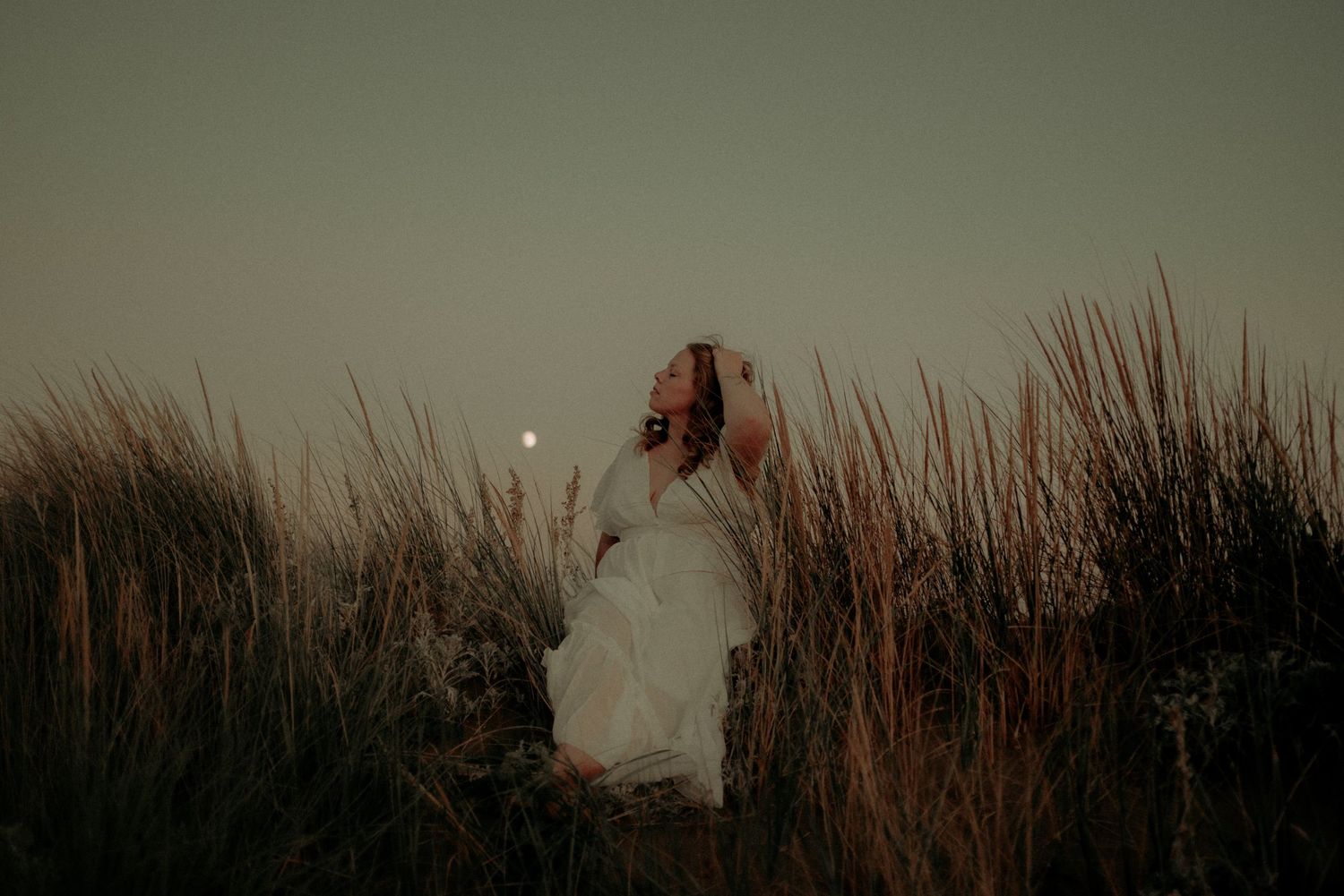 A figure in a white dress stands among tall grass at dusk with a glowing moon in the atmospheric background.