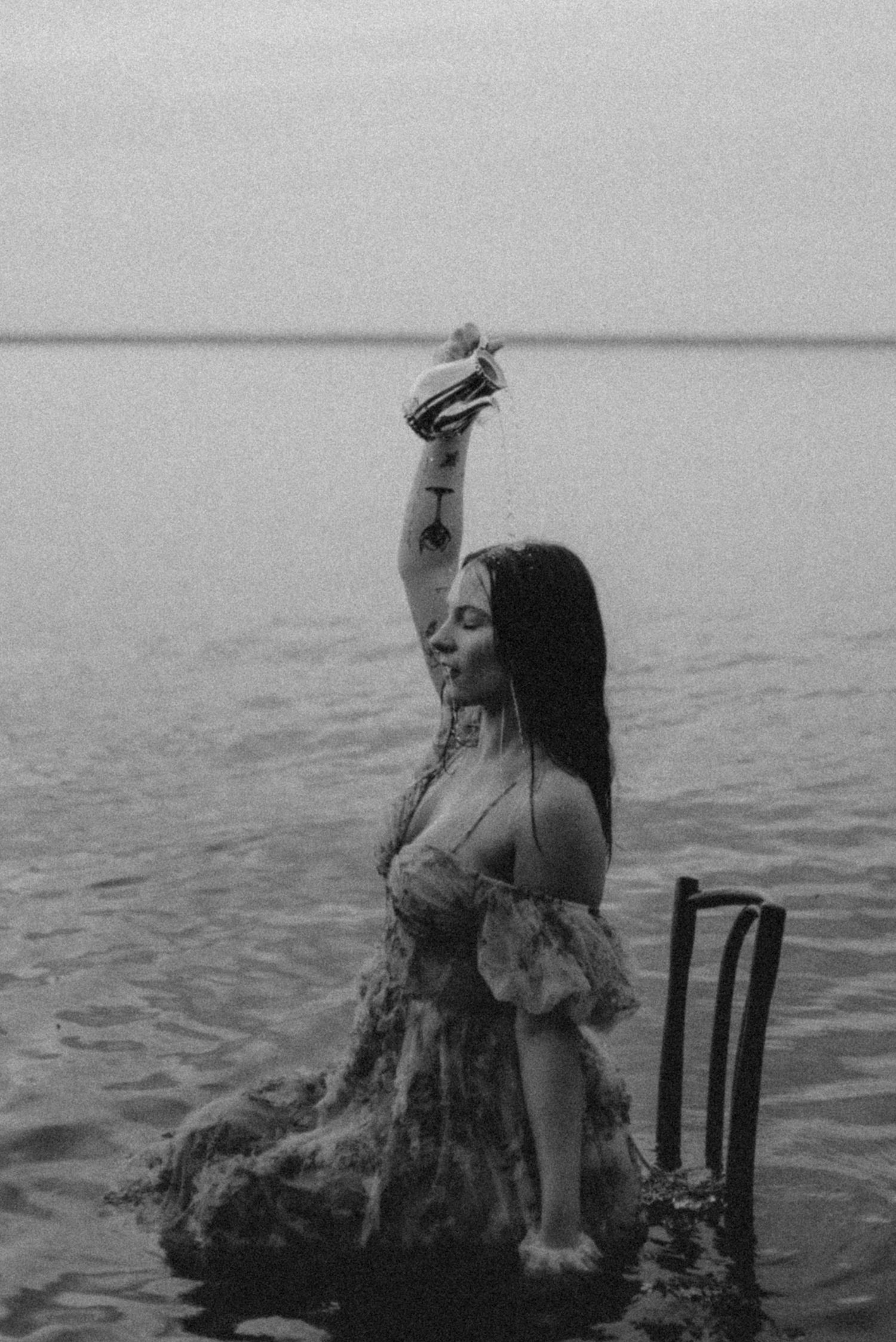 Black and white portrait of a person in vintage dress posed dramatically on chair in water.