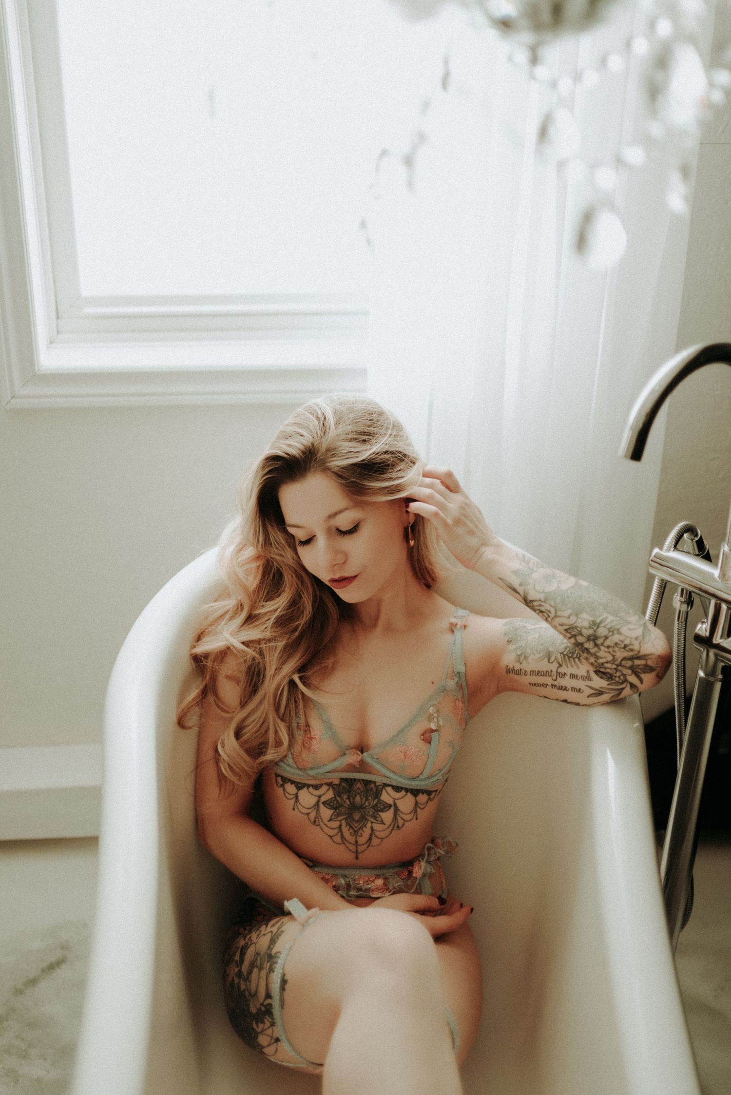 Artistic boudoir photo of a woman with tattoos sitting in a white clawfoot bathtub near a window.
