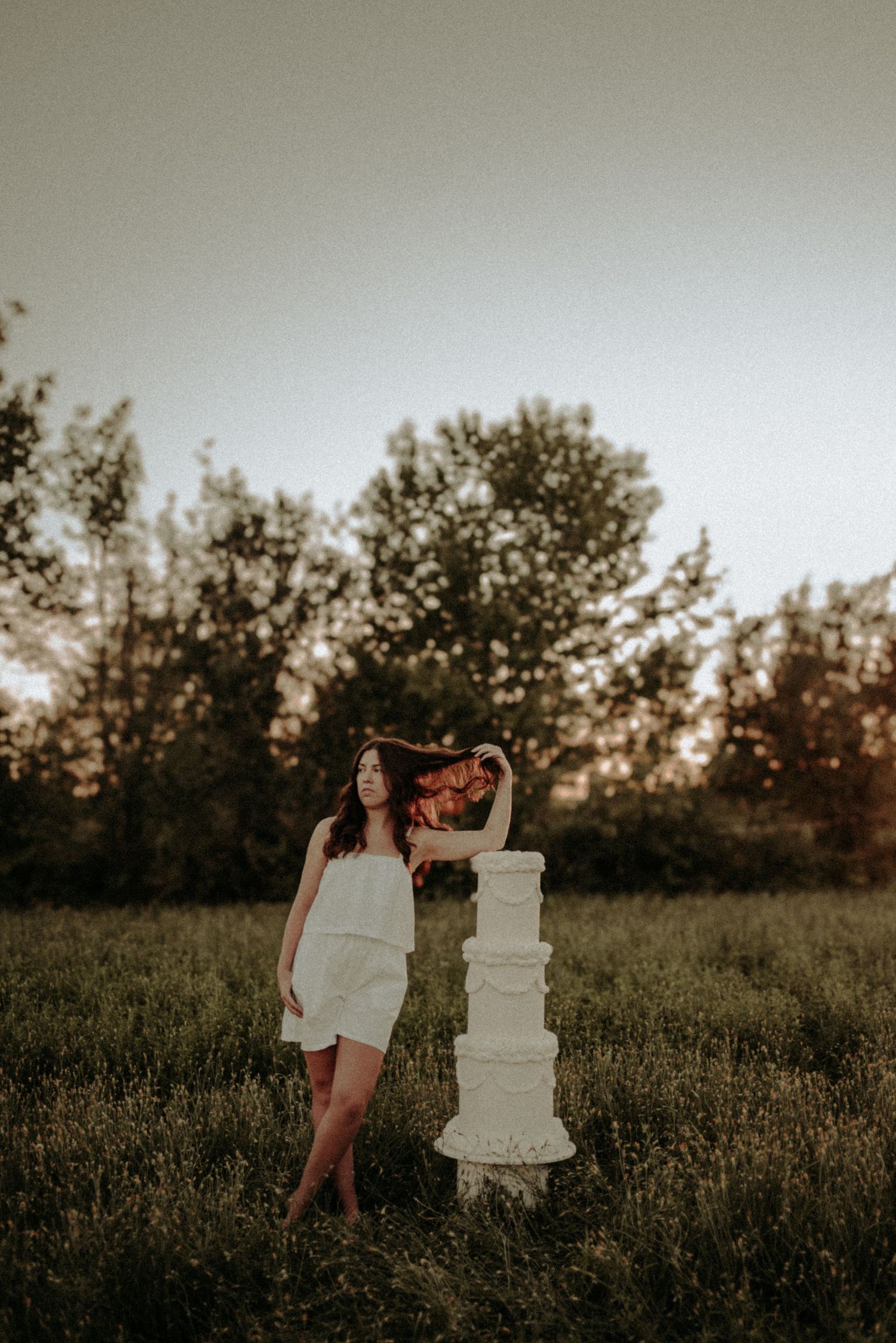 A person in a white dress poses with a decorative white wedding cake in a grassy field at sunset.