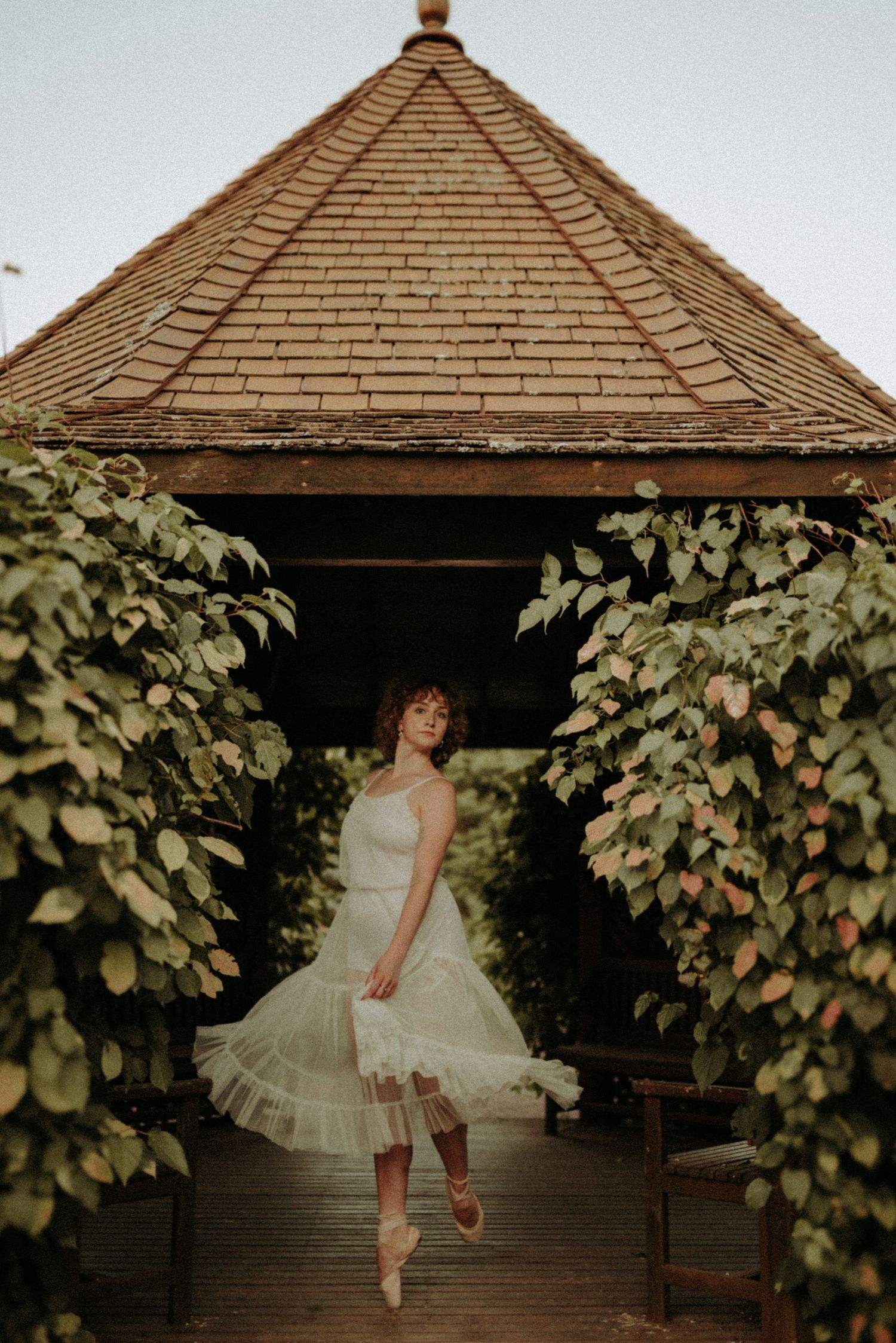 A dancer in a white tulle dress performs graceful ballet poses in front of a rustic gazebo surrounded by green vines.
