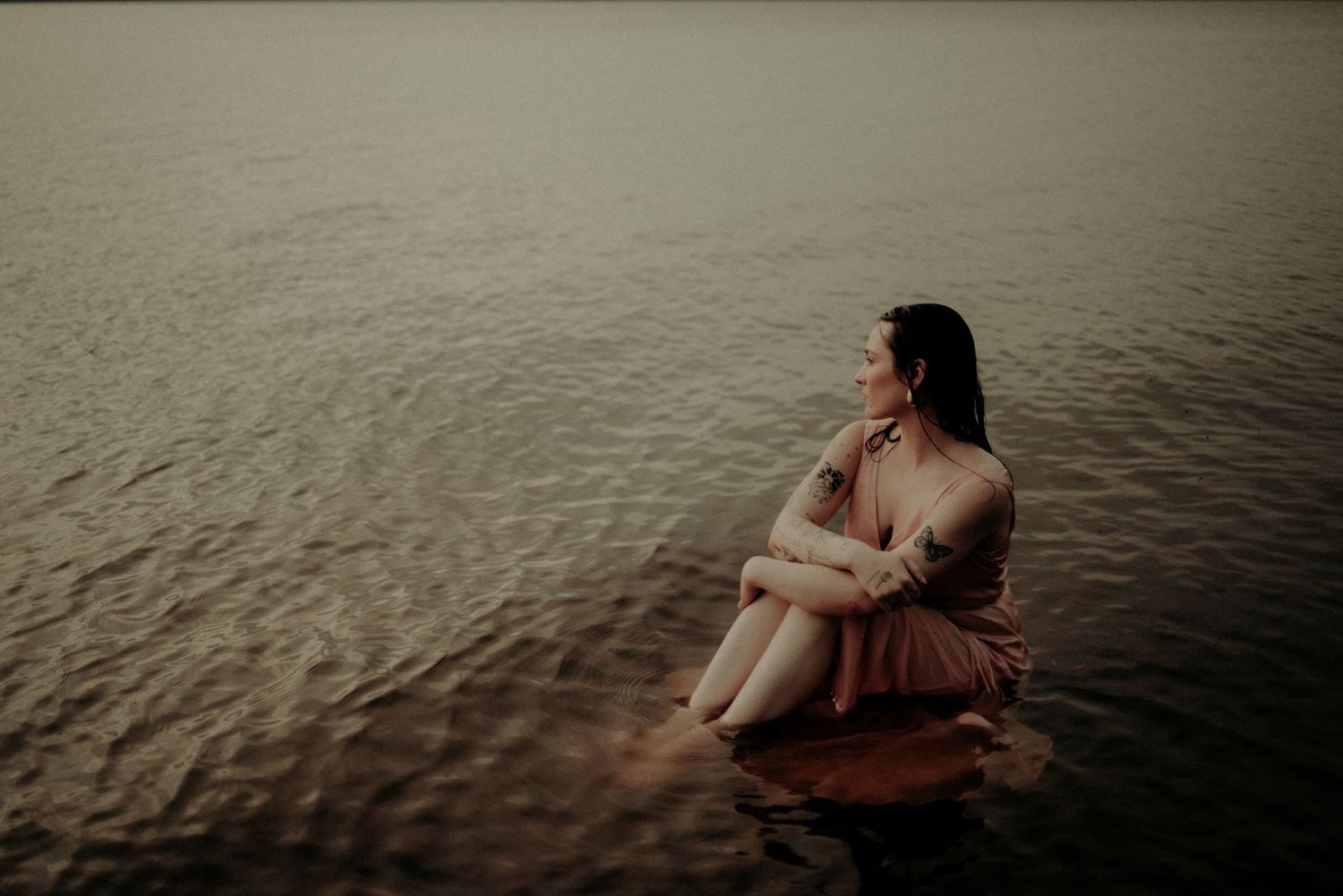 Artistic portrait of a figure in a mauve dress sitting peacefully in still water.