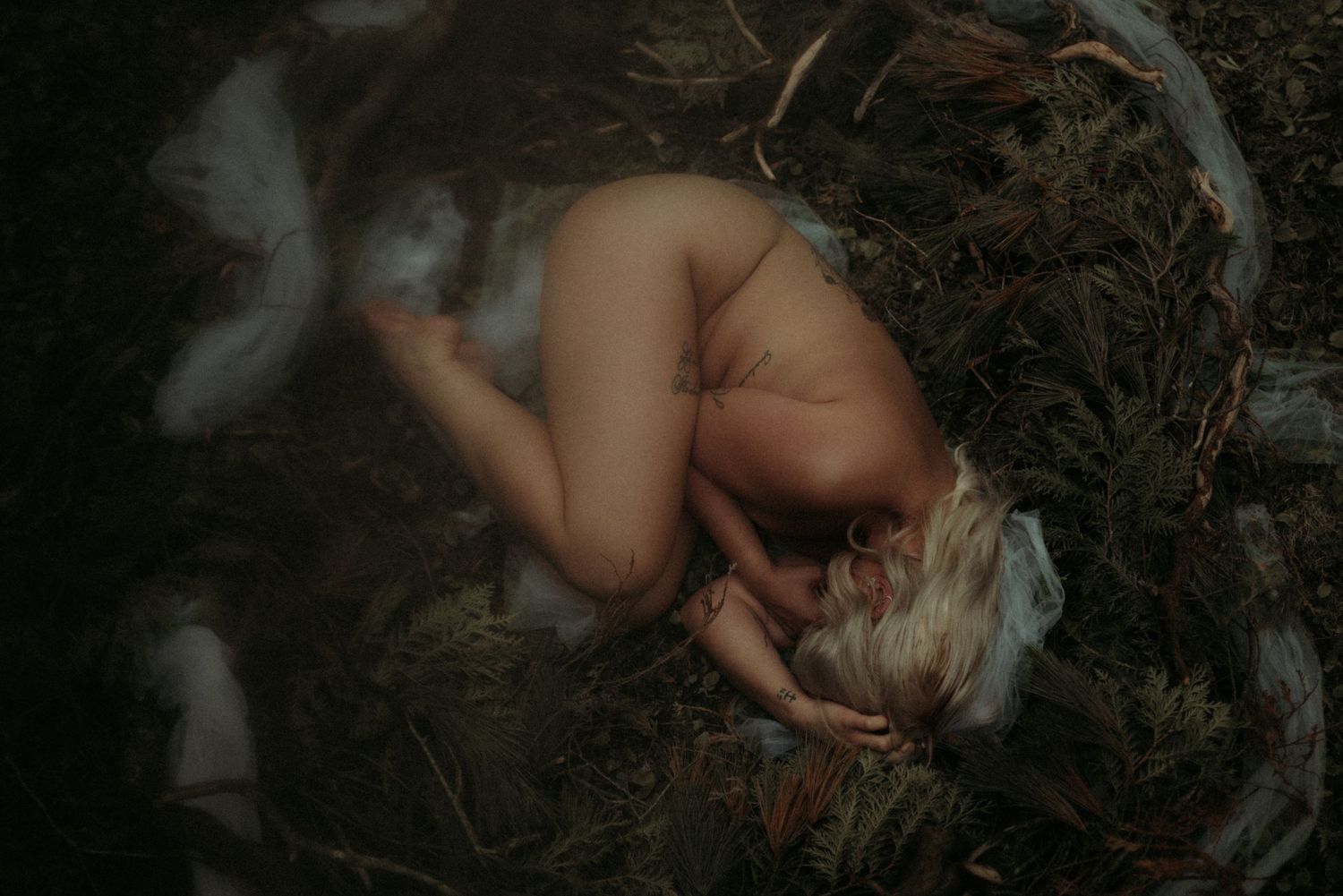 Artistic nude figure curled up on forest floor with ethereal white fabric and natural lighting.