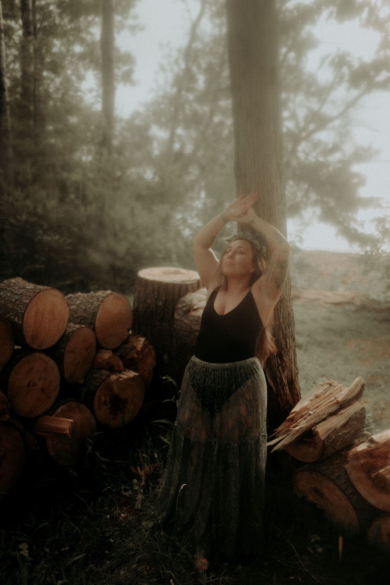 Atmospheric forest portrait with woodpile and stretching pose in muted lighting.