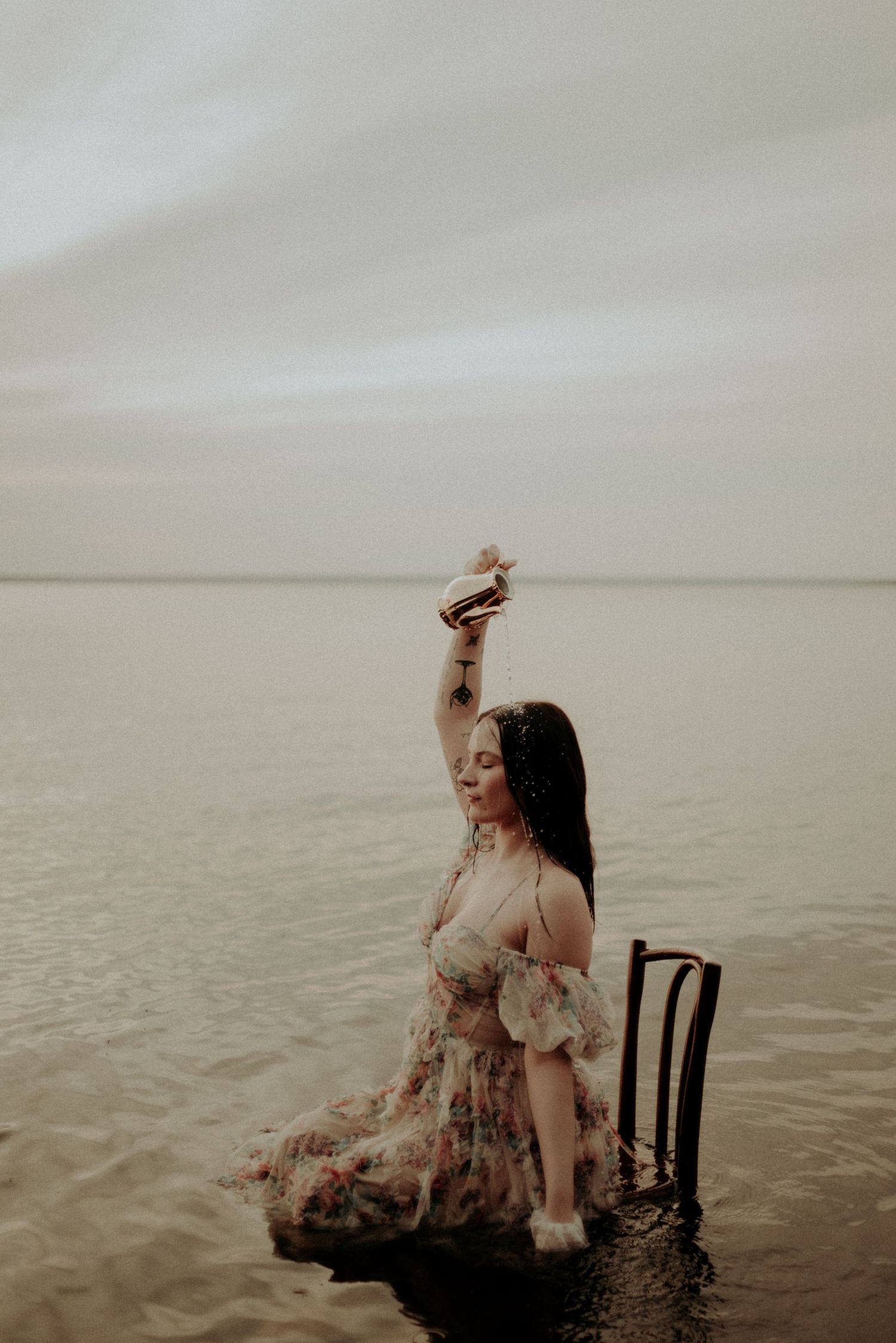 An ethereal water portrait featuring someone in a flowy dress seated on a chair in shallow water.