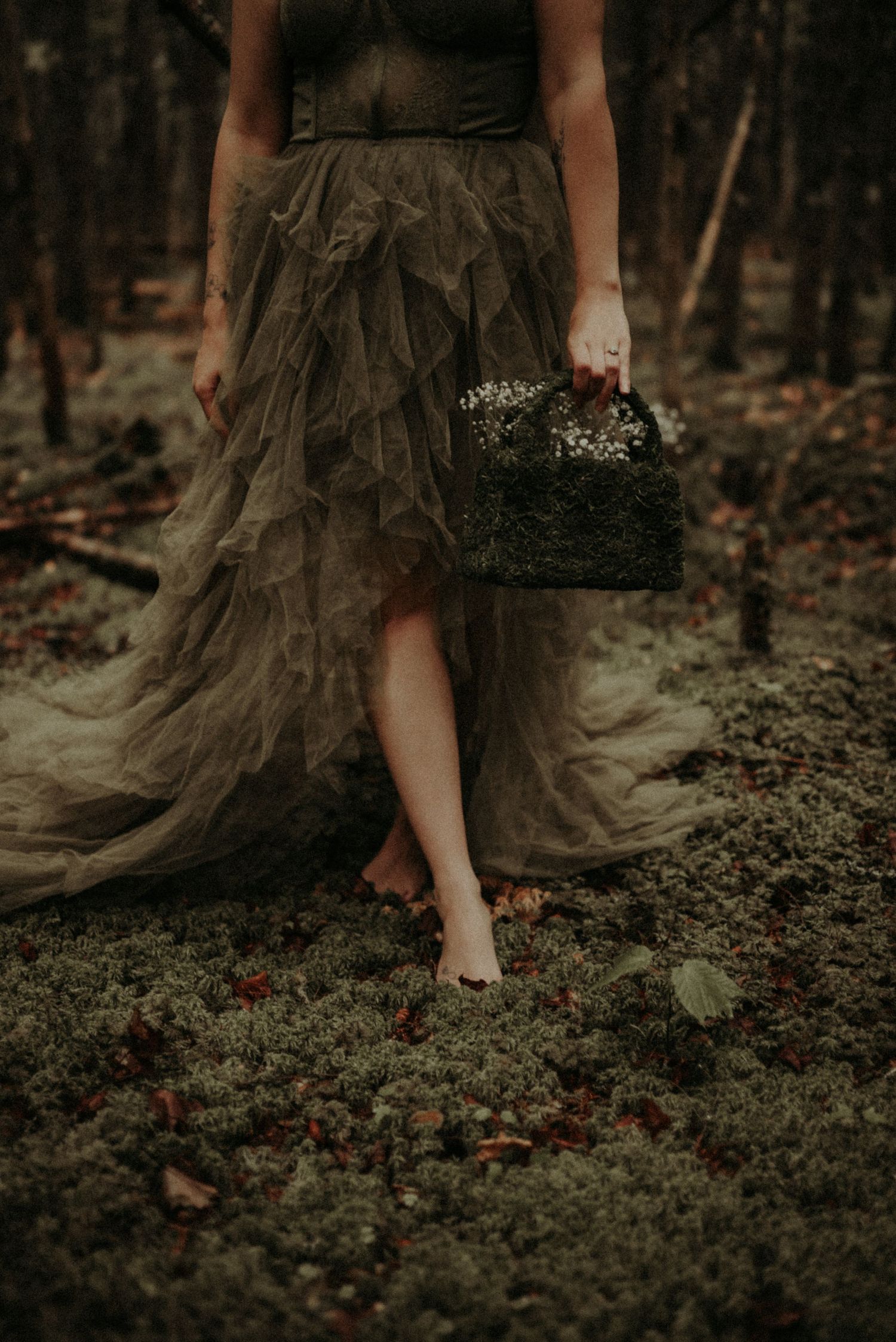 A flowing dark dress brushes against forest floor foliage in a moody woodland setting.