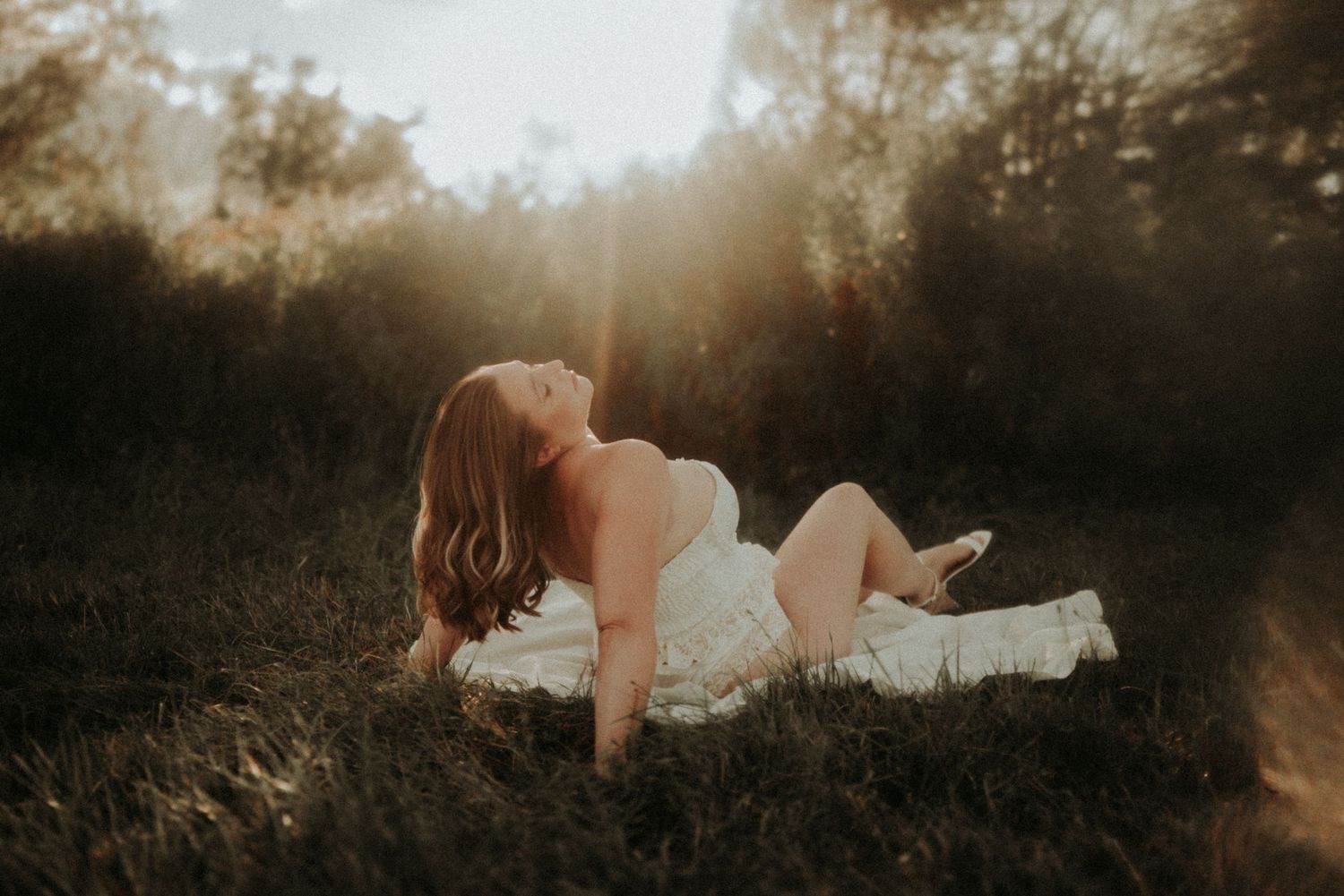 A figure in a white dress reclines in a sunlit meadow during golden hour as sunbeams filter through trees.
