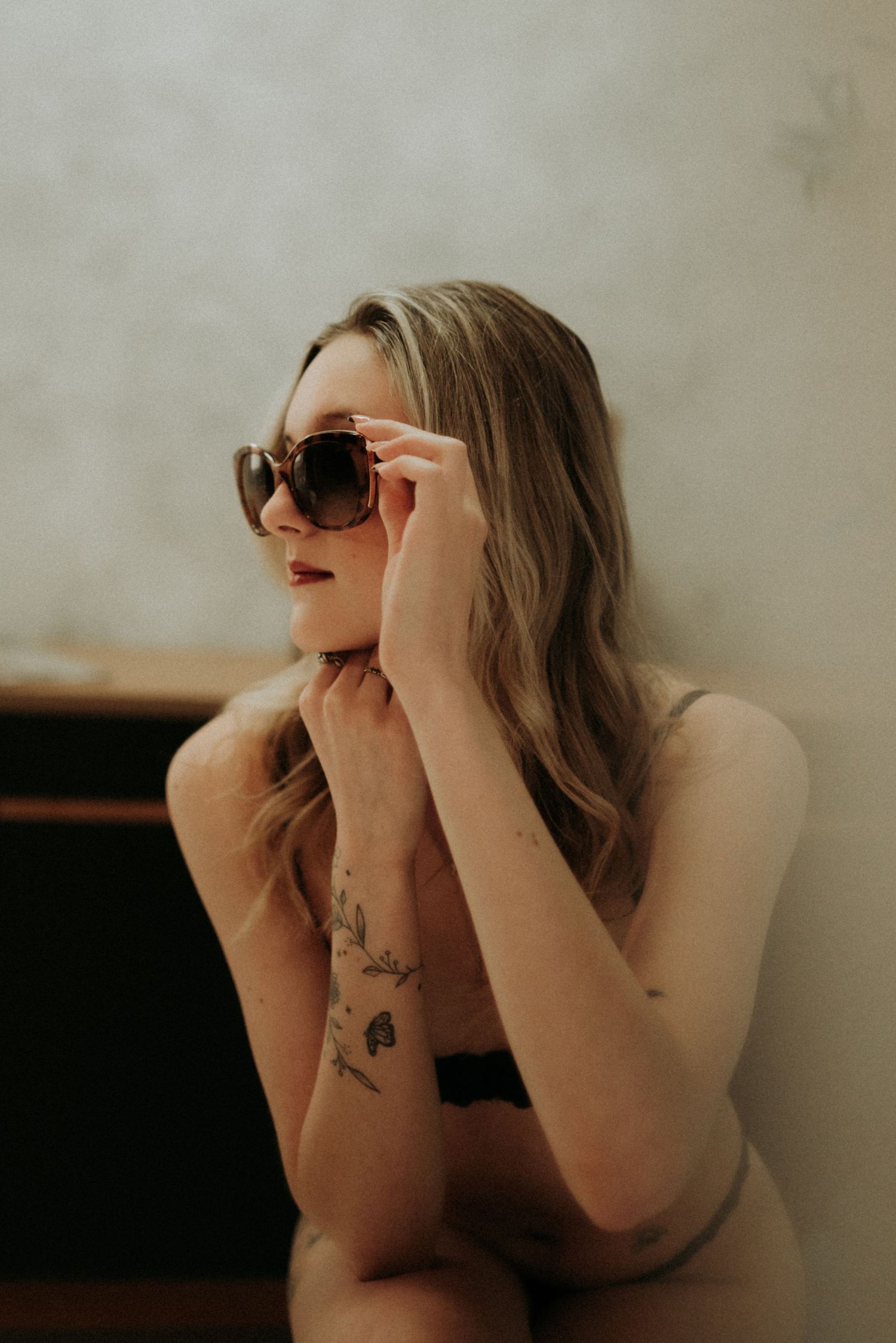 Artistic portrait of person in sunglasses with visible arm tattoos posing against neutral background in warm lighting.