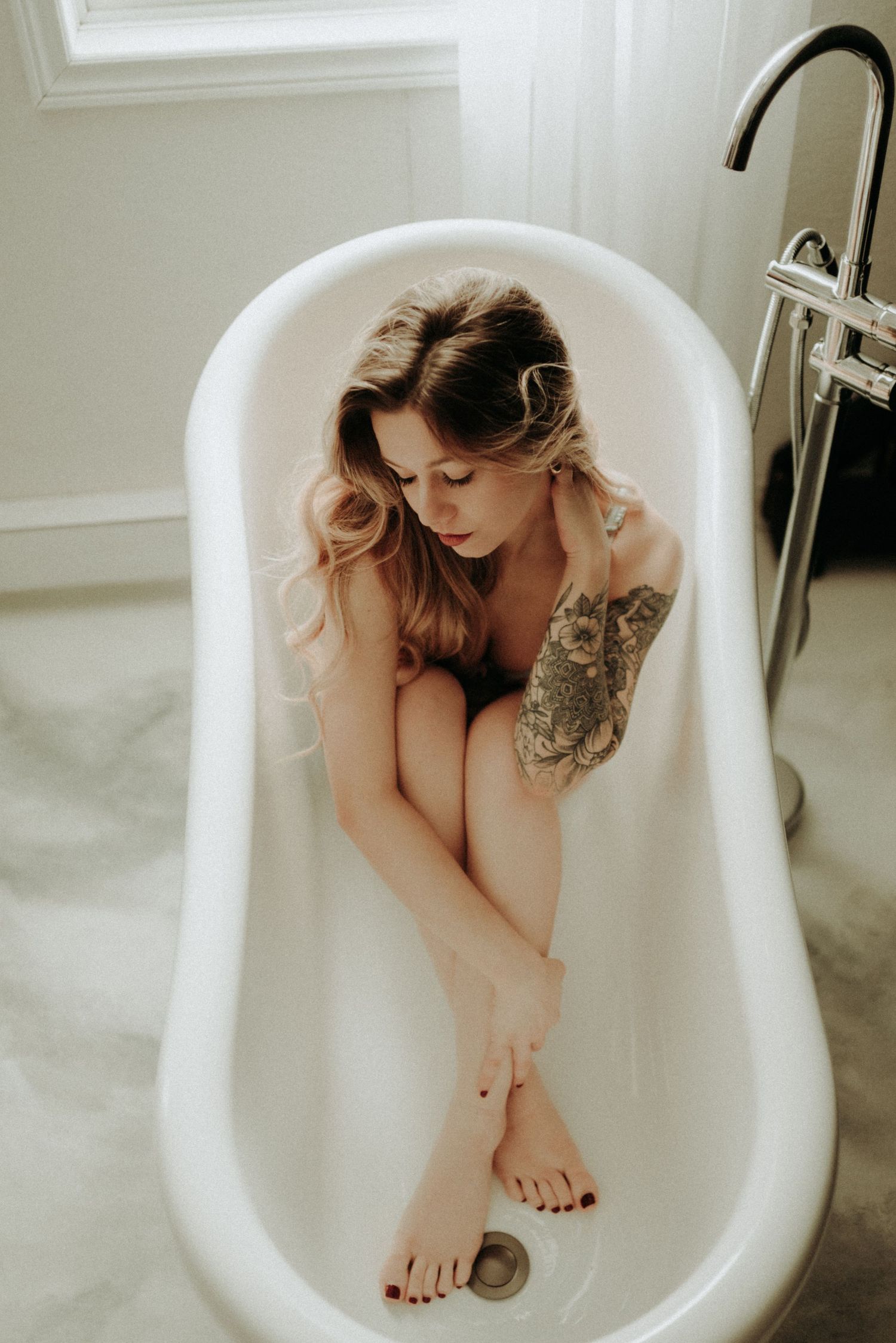 Artistic boudoir photo of a woman sitting contemplatively in a white freestanding bathtub.