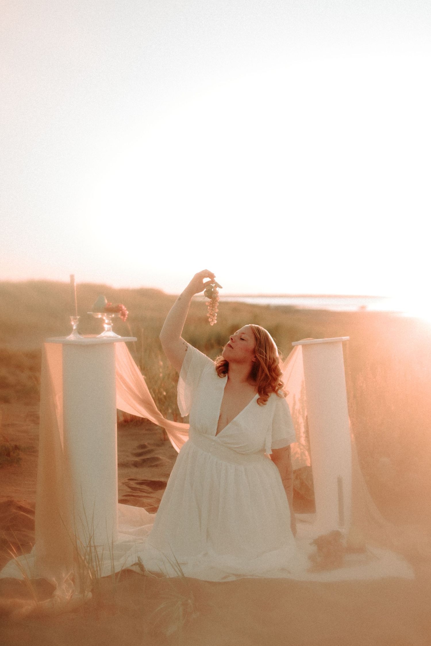 A flowing white dress billows in the wind against a sunset backdrop with white pillars on a sandy beach.