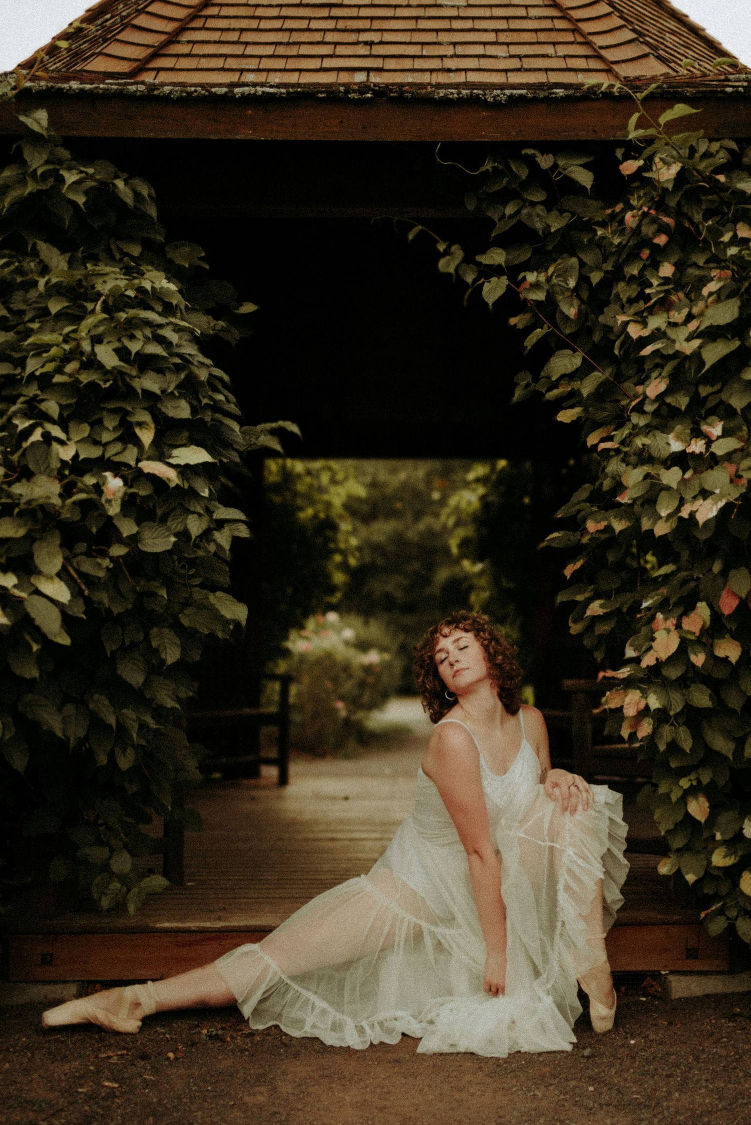A figure in a vintage white dress reclines gracefully on a rustic porch with climbing vines and dappled sunlight.