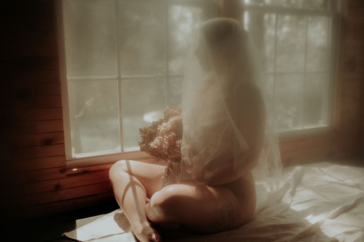 Dreamy boudoir photograph of someone sitting by a window in soft morning light.