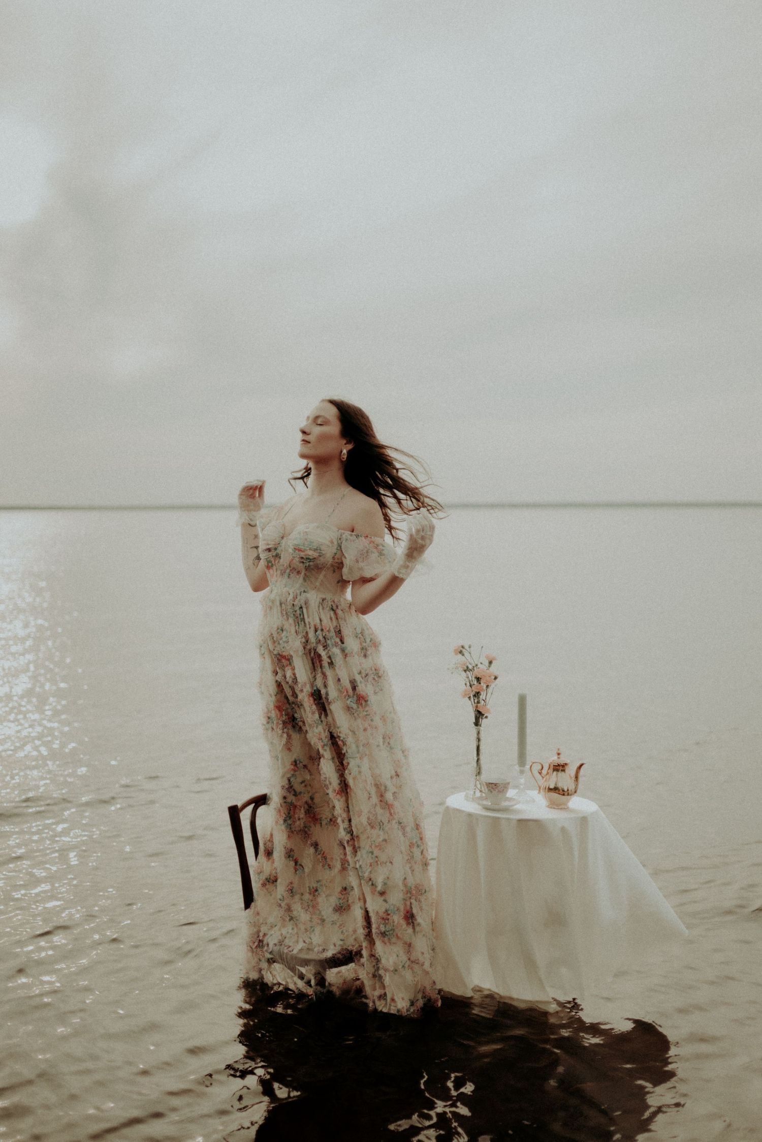 Ethereal portrait of a floral dress in shallow water with a white table setting and delicate flowers against a misty horizon.