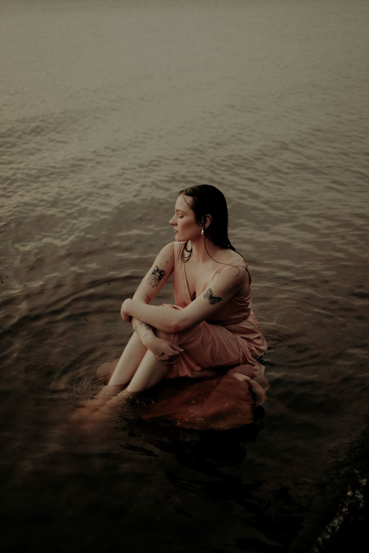 A dramatic moody photo of someone sitting in shallow water wearing a flowy pink dress at sunset.
