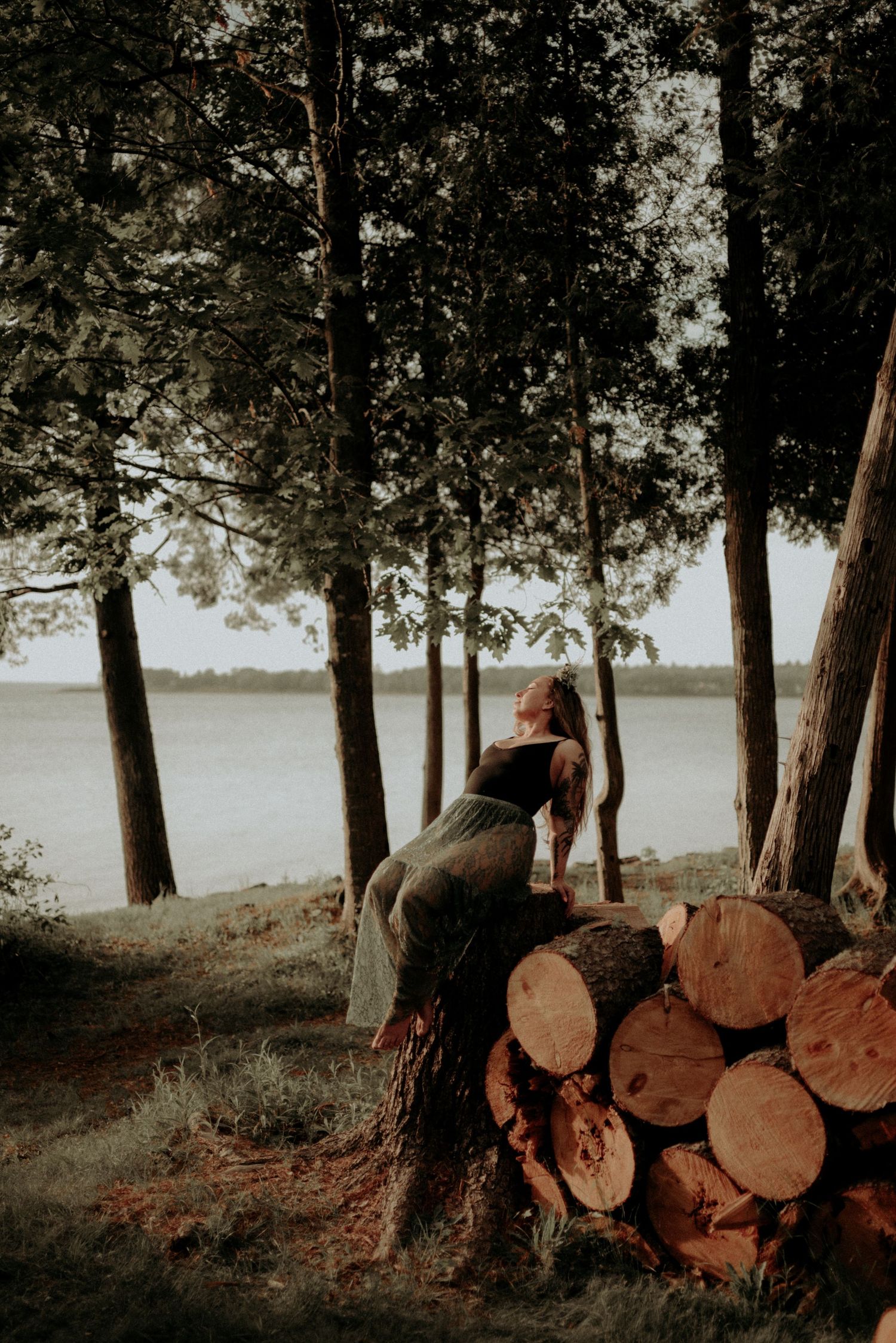 Figure posed against stacked logs in waterfront forest scene at dusk.
