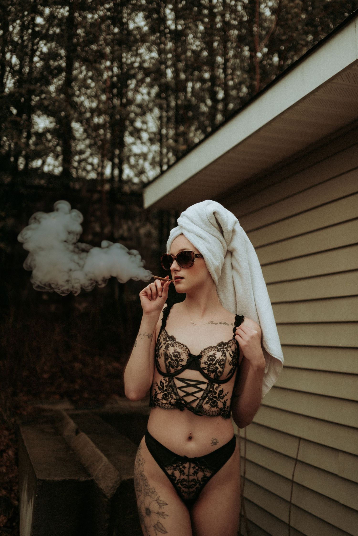 Artistic boudoir portrait of figure in black lace lingerie and head wrap smoking against house exterior.